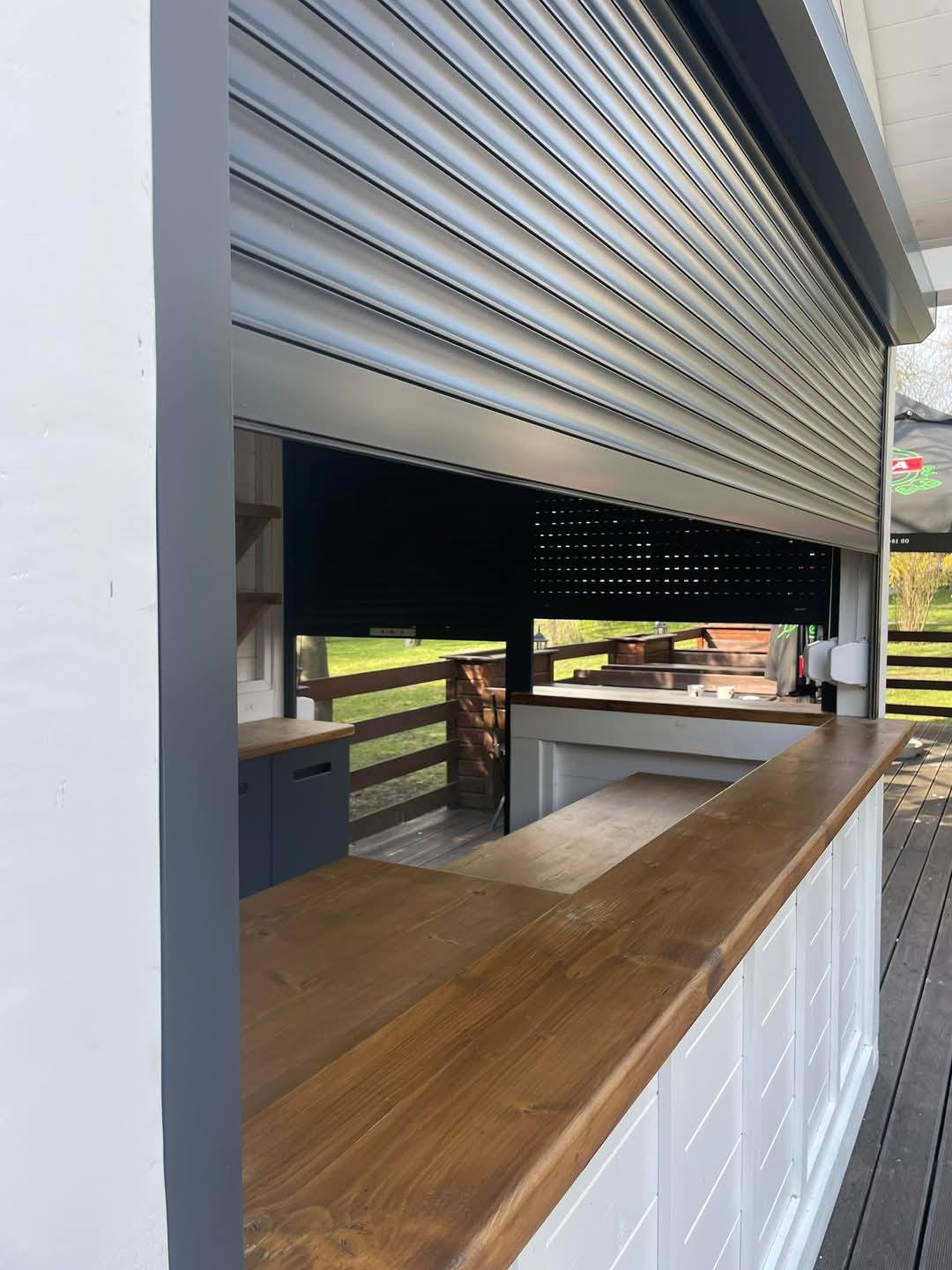 Częściowo opuszczona, szara przesłona okienna z lamelami, widoczna z zewnątrz budynku. Drewniany blat i białe panele poniżej. Widok na zewnątrz przez otwartą przestrzeń.