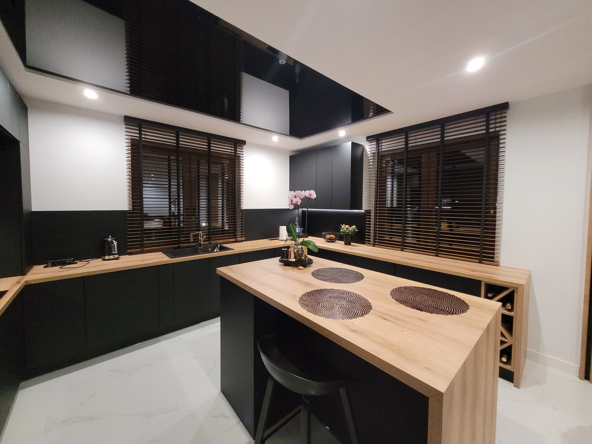 Nowoczesna kuchnia z drewnianymi żaluzjami w oknach. Czarne szafki i jasny blat tworzą kontrast. Widoczne detale wystroju i oświetlenie.