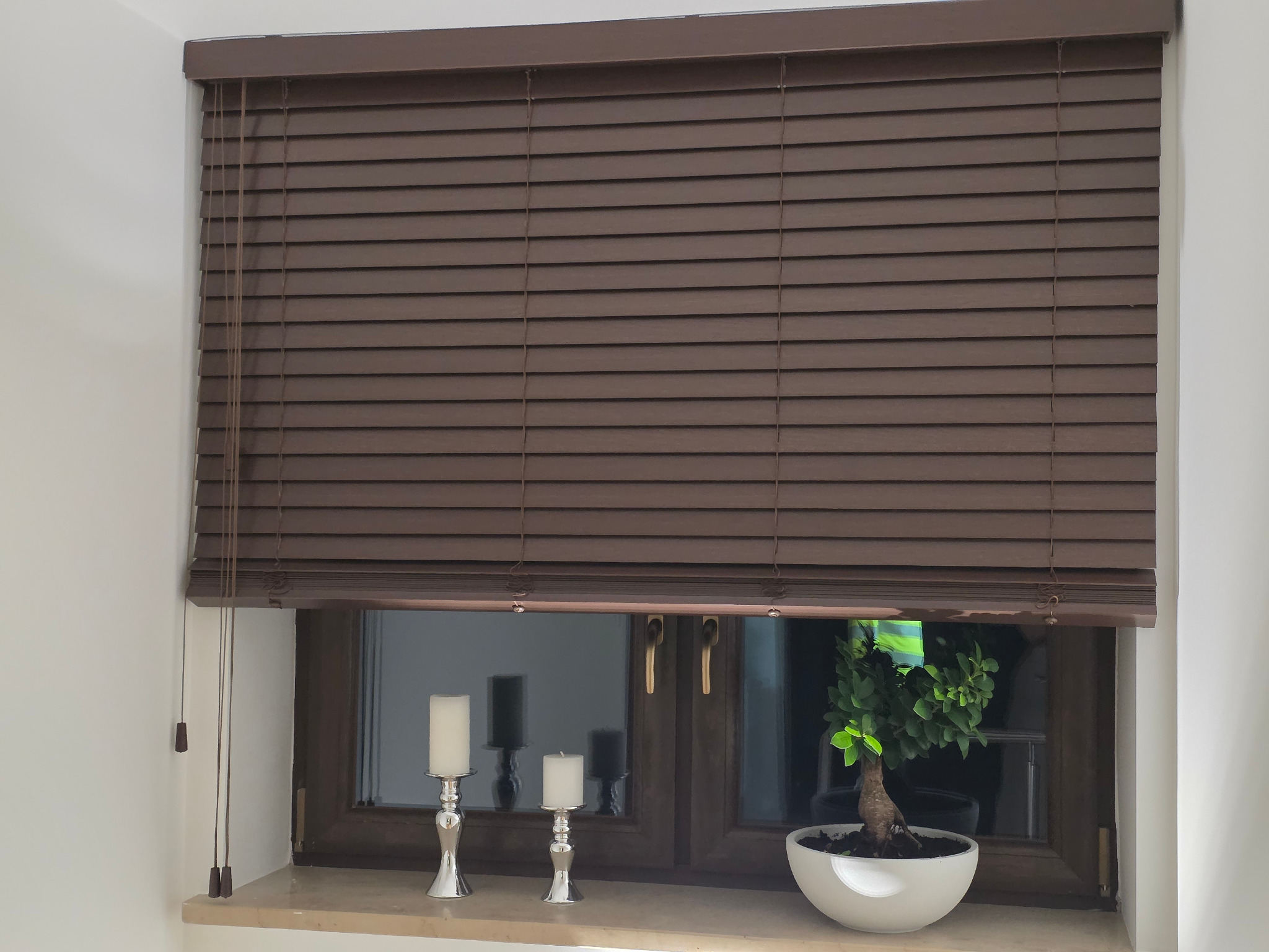 Brązowe żaluzje drewniane zamontowane w oknie, z widocznymi sznurkami do regulacji. Na parapecie dekoracje: świeczniki i bonsai. Ujęcie z bliska, wnętrze.
