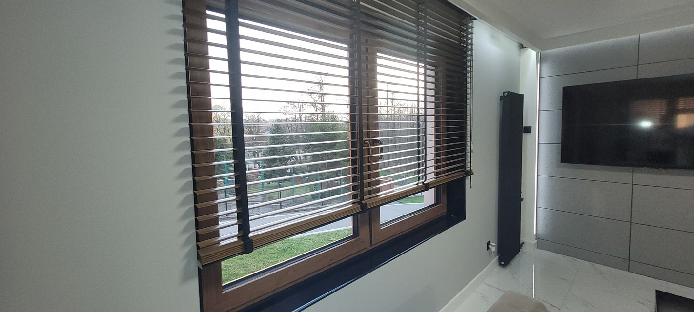 Drewniane żaluzje poziome w oknie z widokiem na zieleń. Ciepłe odcienie drewna kontrastują z jasnymi ścianami i nowoczesnym wnętrzem. Żaluzje częściowo zasłonięte.