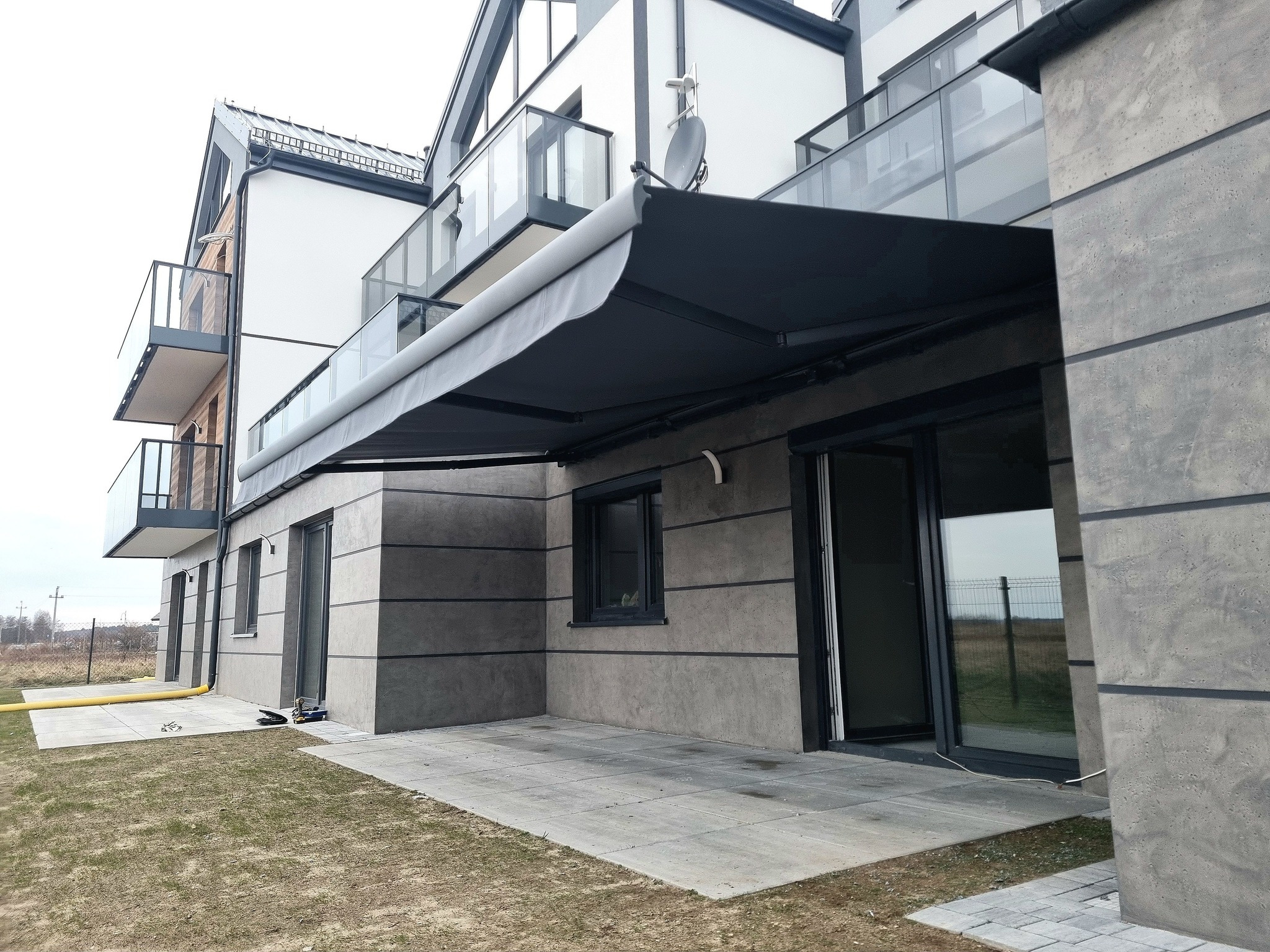 Zewnętrzna, grafitowa markiza tarasowa zamontowana przy nowoczesnym budynku z balkonami i szklanymi balustradami. Widok na taras z szarych płyt.