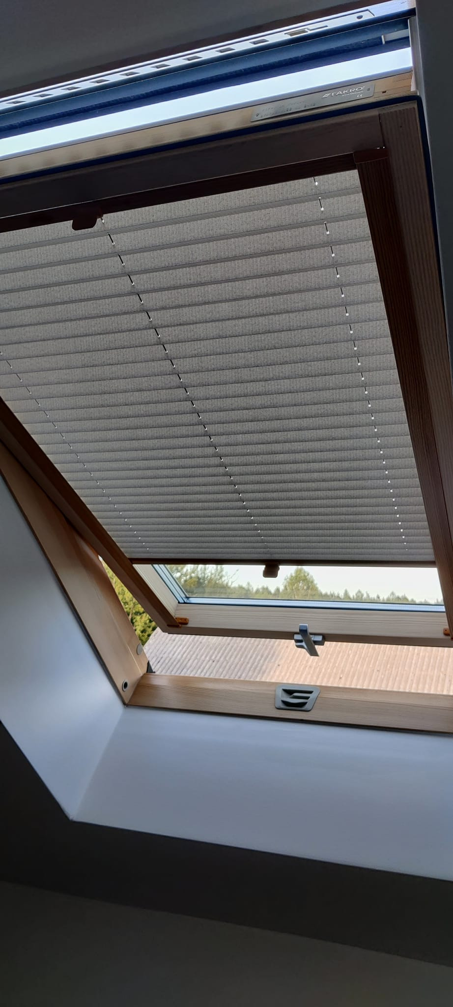 Szara, plisowana roleta okienna marki Fakro, zamontowana w oknie dachowym z drewnianą ramą. Widok od wewnątrz, na zewnątrz widać zieleń drzew.
