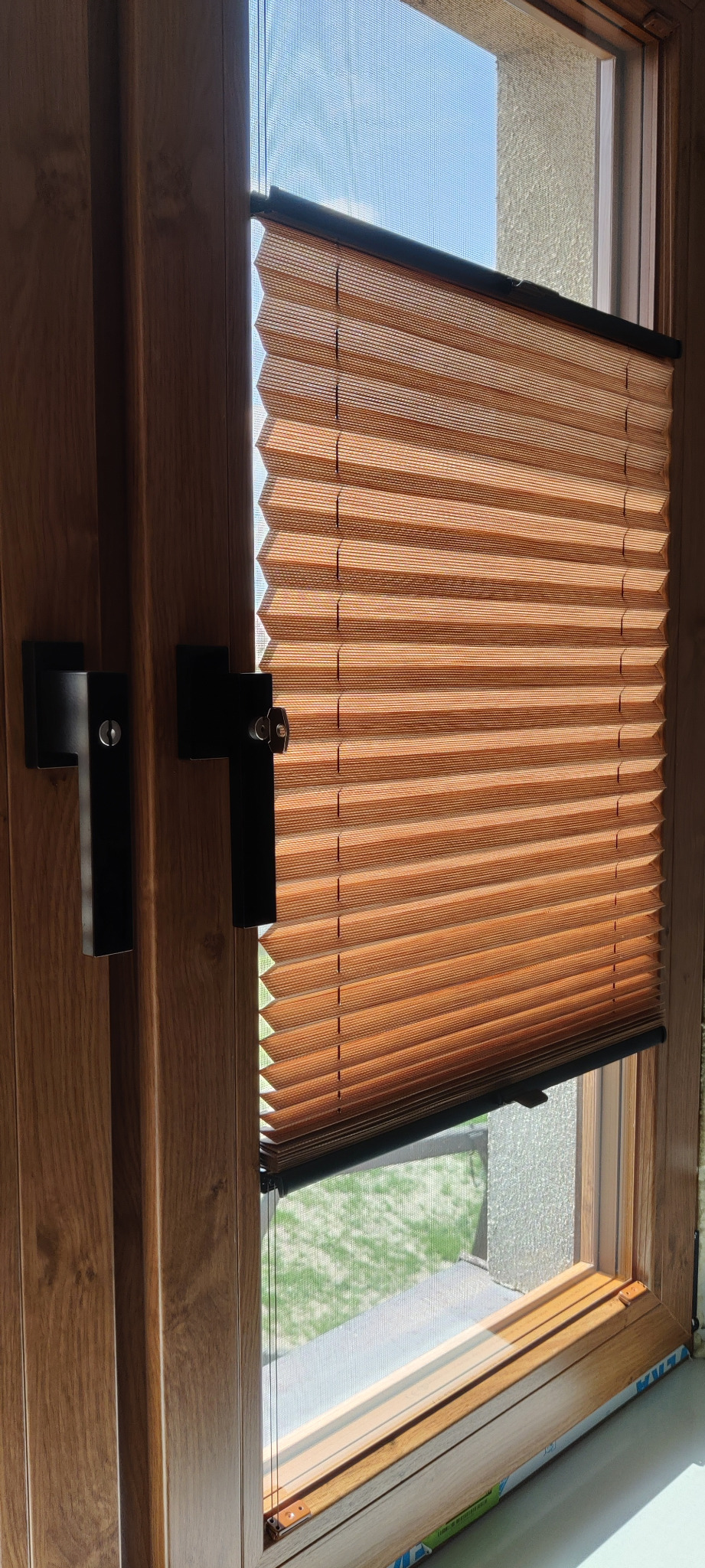 Brązowa plisa okienna zamontowana w drewnianym oknie, widok z bliska na plisę z czarnymi elementami montażowymi i uchwytem okiennym. Widok na zewnątrz przez okno.