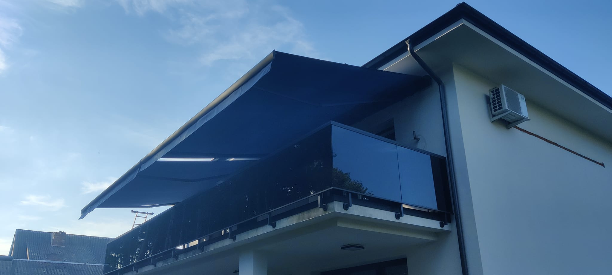 Elegancka, rozłożona markiza w kolorze granatowym nad szklanym balkonem nowoczesnego domu, widziana z dołu pod kątem. Błękitne niebo w tle.