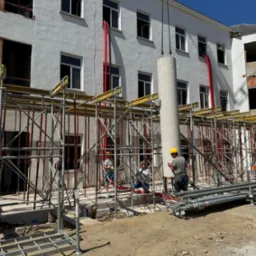 Montaż prefabrykowanego słupa na budowie budynku z białymi ścianami. Widoczne rusztowania, pracownicy w kaskach i czerwone rury wentylacyjne.