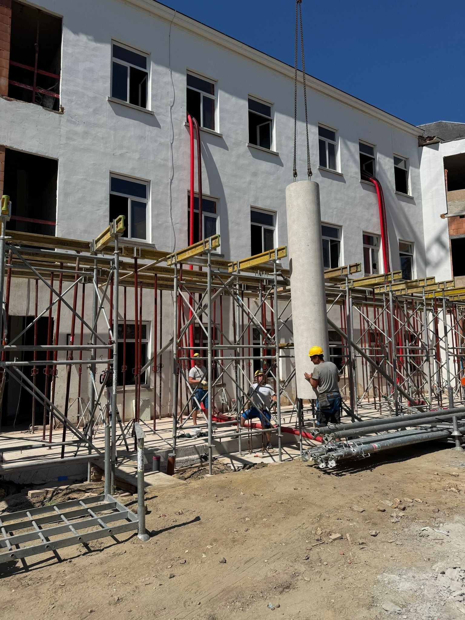 Montaż prefabrykowanego słupa na budowie budynku z białymi ścianami. Widoczne rusztowania, pracownicy w kaskach i czerwone rury wentylacyjne.
