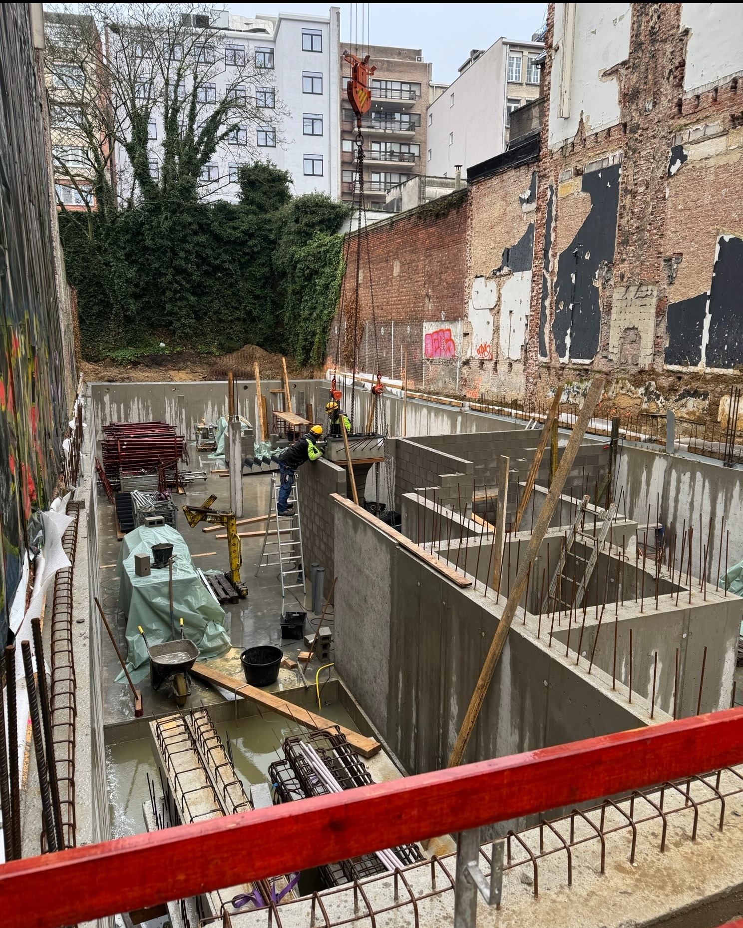 Zalane fundamenty budynku z widocznym zbrojeniem i betonowymi ścianami. Na budowie pracują robotnicy w kaskach. W tle budynki mieszkalne i zieleń.