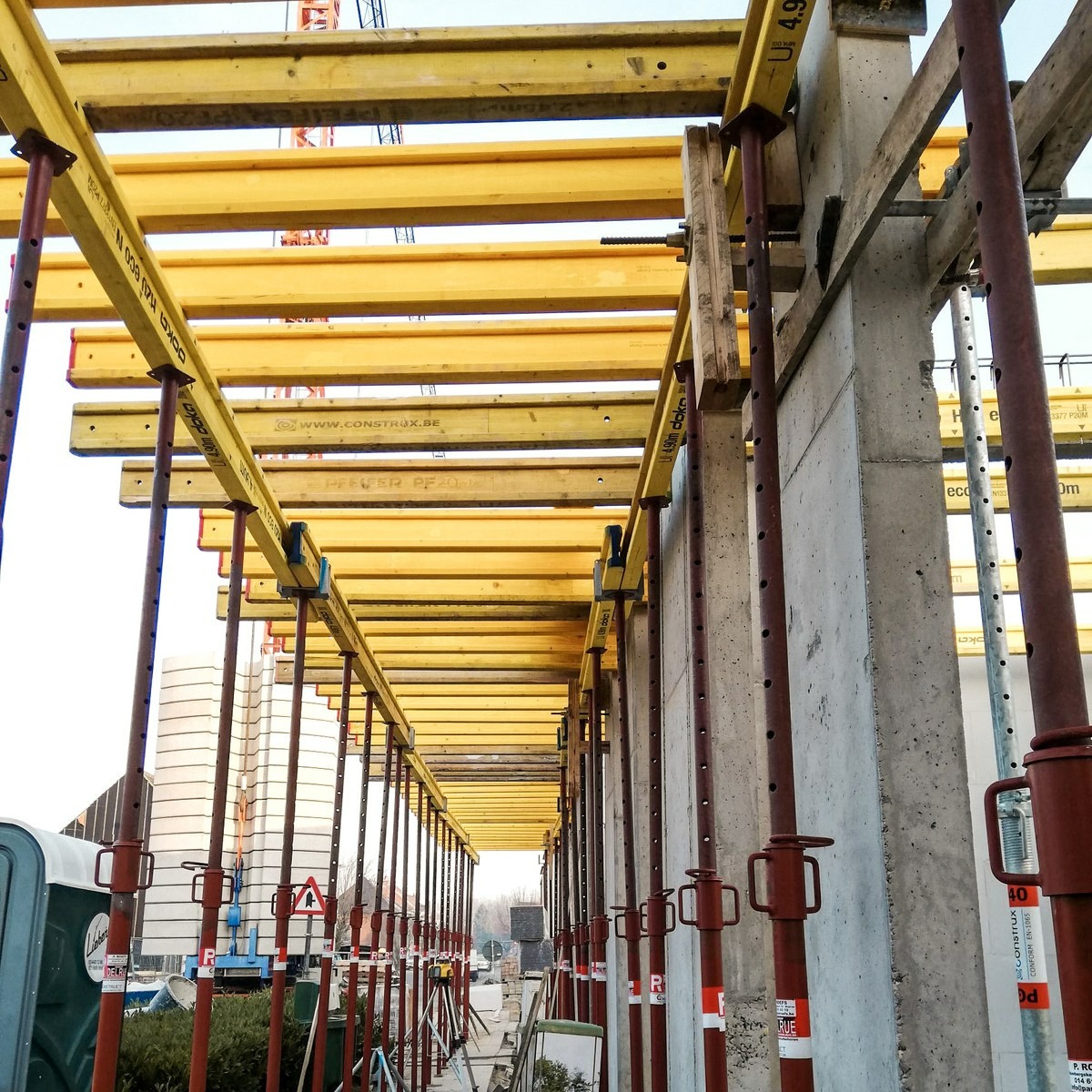Szare, betonowe słupy podtrzymują żółte, drewniane belki stropowe na budowie. Widoczne podpory i elementy konstrukcyjne. W tle budynki i znaki drogowe.