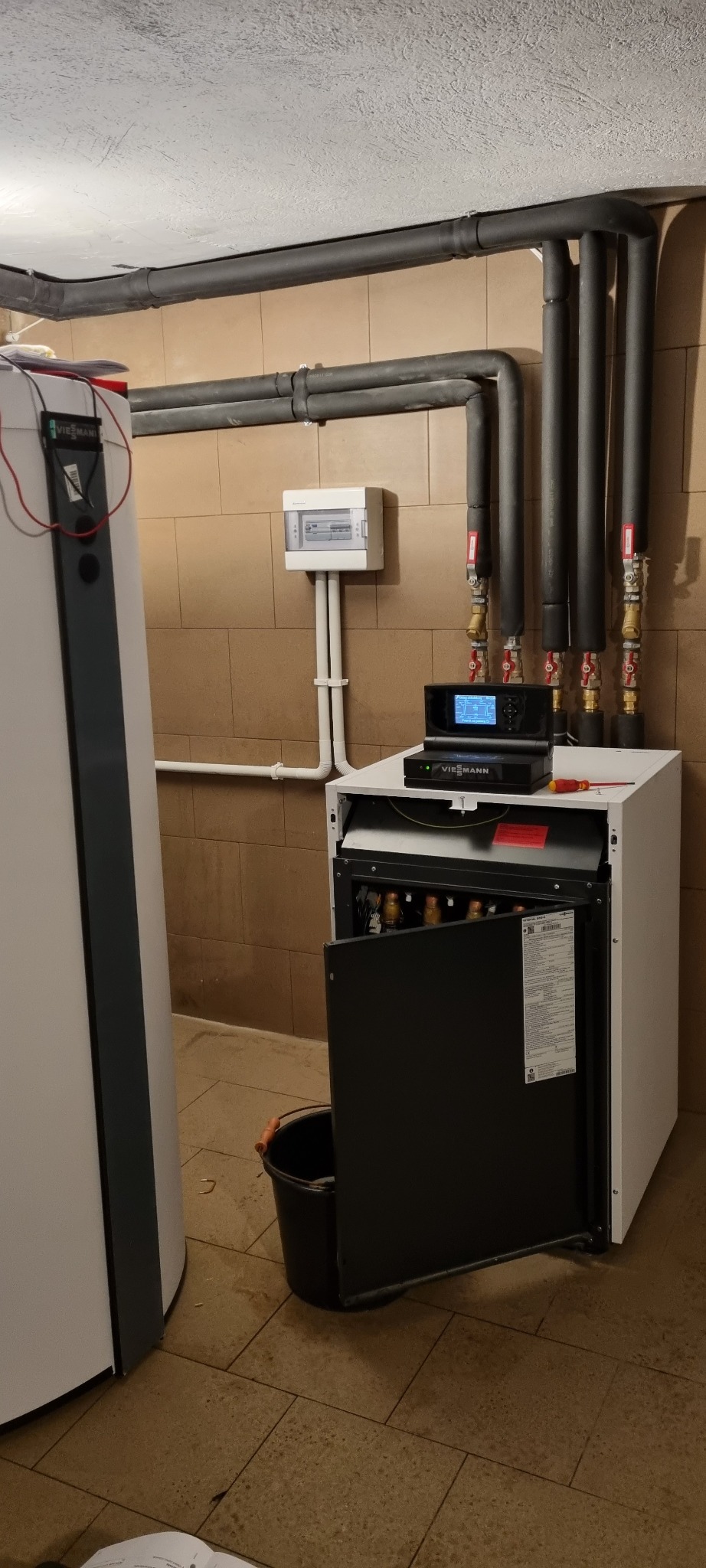 Otwarty piec Viessmann w kotłowni, widoczne rury instalacji grzewczej, czarny sterownik na piecu i skrzynka elektryczna na ścianie.