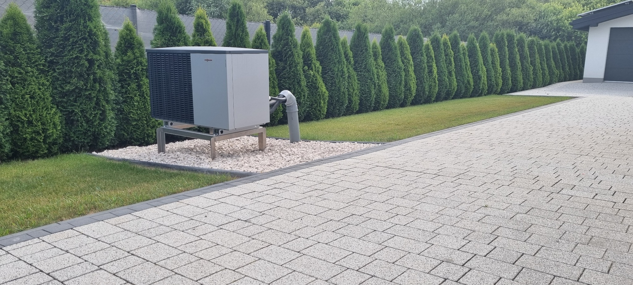 Szara pompa ciepła Viessmann na metalowym stelażu, otoczona białym żwirem, na tle rzędu tuj i kostki brukowej. Instalacja na zewnątrz budynku.