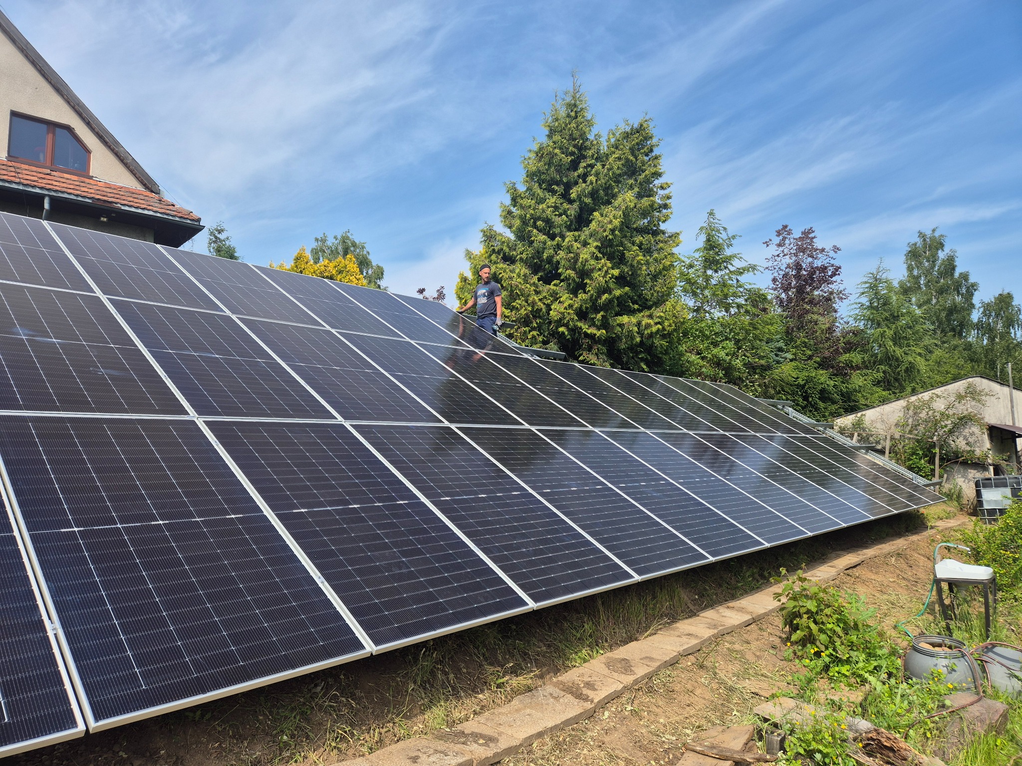 Instalacja fotowoltaiczna na dachu domu jednorodzinnego, widok z dołu, monter na dachu, błękitne niebo w tle, widoczne drzewa i zieleń.