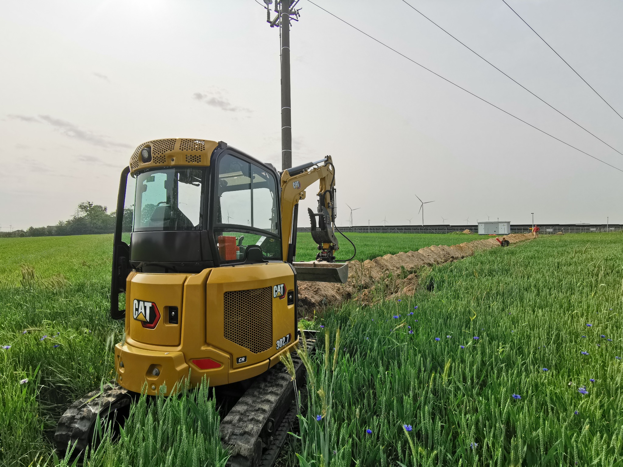 Żółta mini koparka CAT 302.7 w zielonym polu, w tle wykopany rów, słup elektryczny i turbiny wiatrowe. Praca w toku, z bliska widoczne detale maszyny.