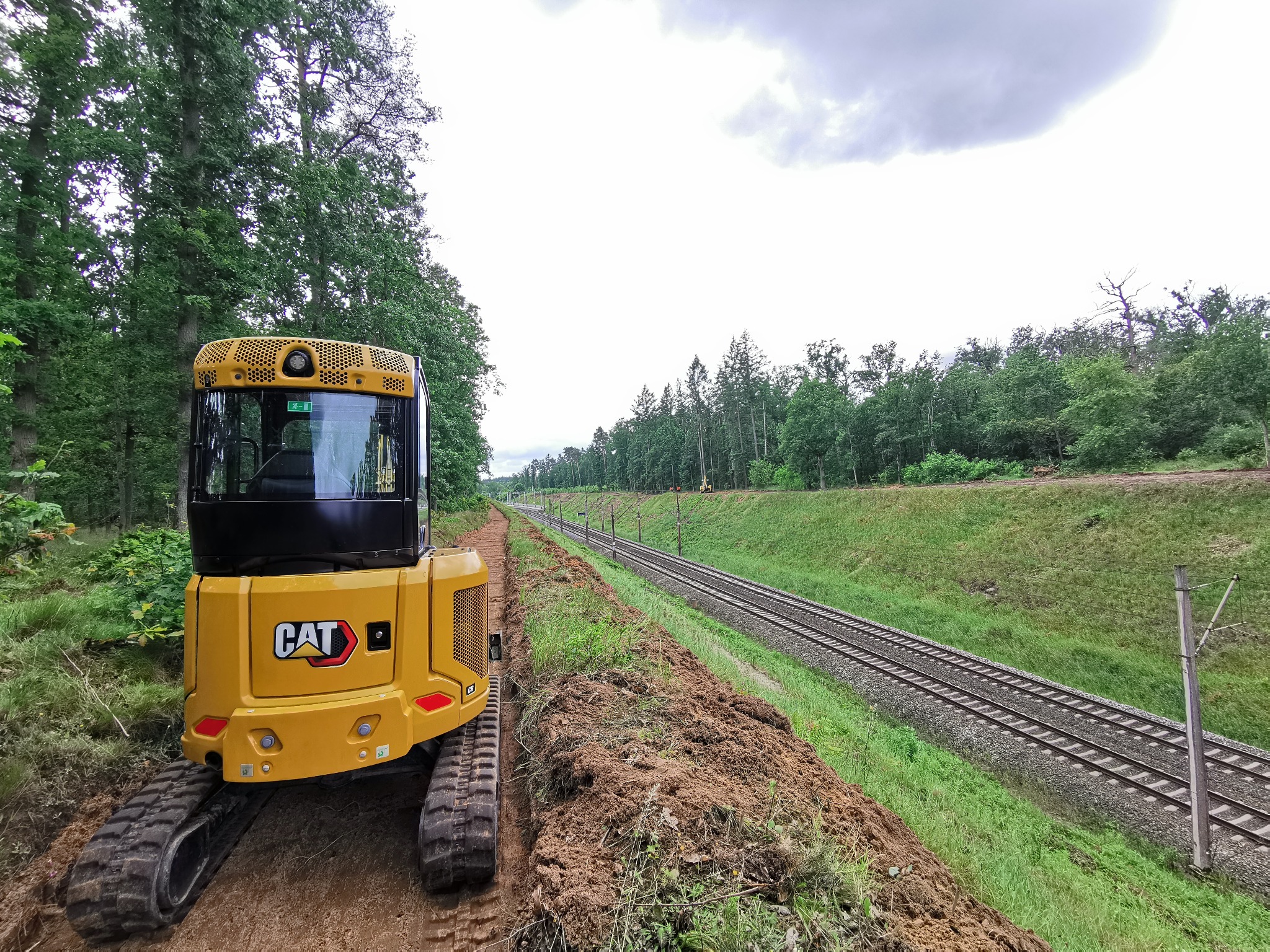 Żółta mini koparka CAT na gąsienicach, obok torów kolejowych. Wykopany rów wzdłuż torów, zieleń w tle. Praca ziemna w trakcie realizacji, widok z tyłu maszyny.