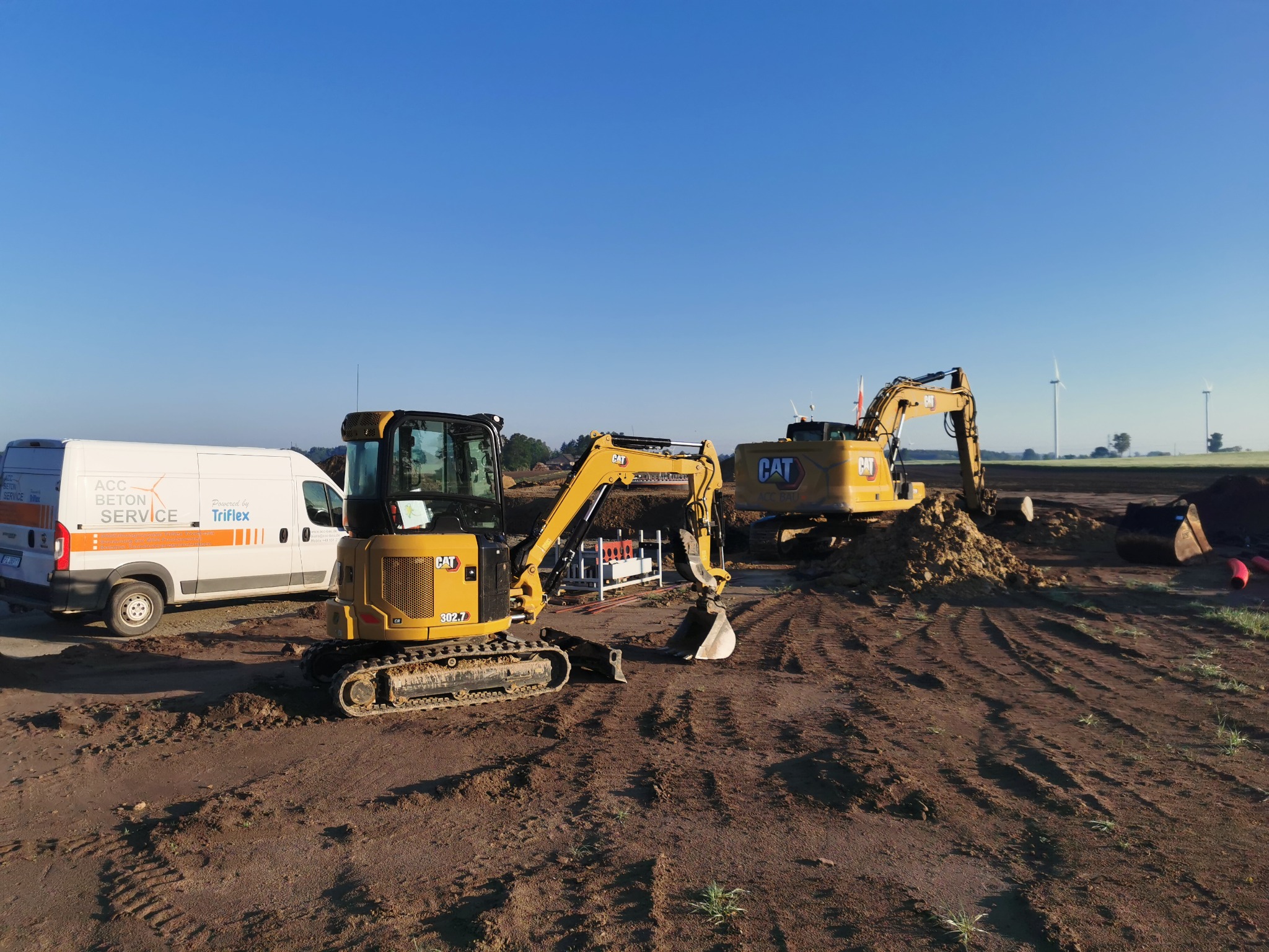 Dwa żółte mini-koparki CAT na placu budowy, z widocznym vanem ACC Beton Service w tle i turbinami wiatrowymi na horyzoncie, w słoneczny dzień.