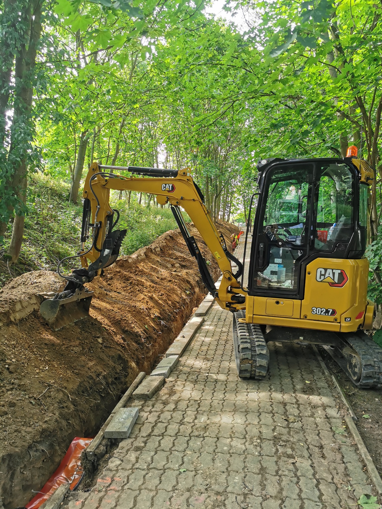 Żółta mini koparka CAT 302.7 wykonuje wąski wykop wzdłuż chodnika z kostki, otoczona zielenią drzew. W wykopie widoczna pomarańczowa folia.