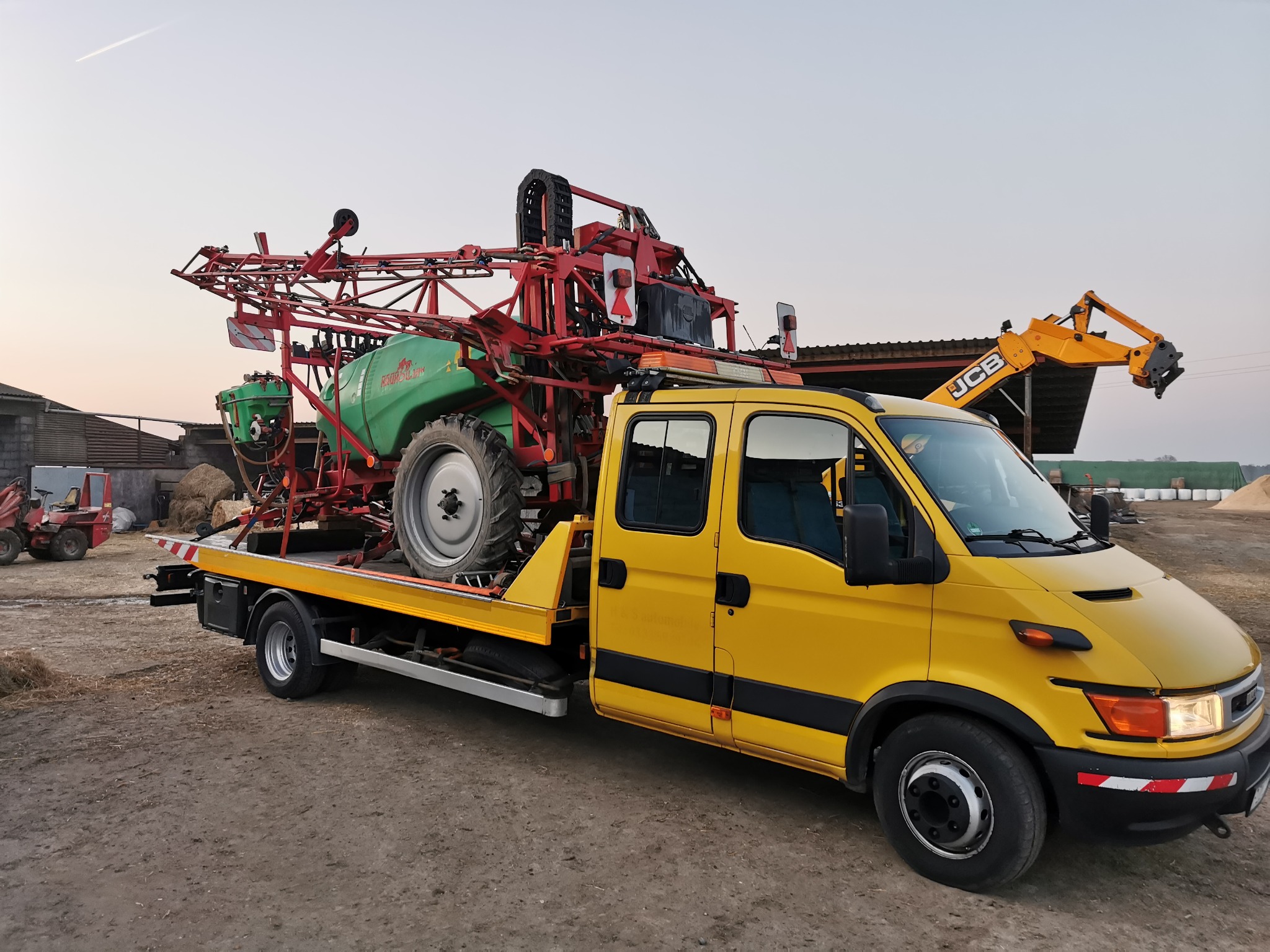 Żółta laweta przewozi zielony opryskiwacz rolniczy marki Holder, z czerwonymi elementami konstrukcyjnymi. W tle budynki gospodarcze i koparka JCB.