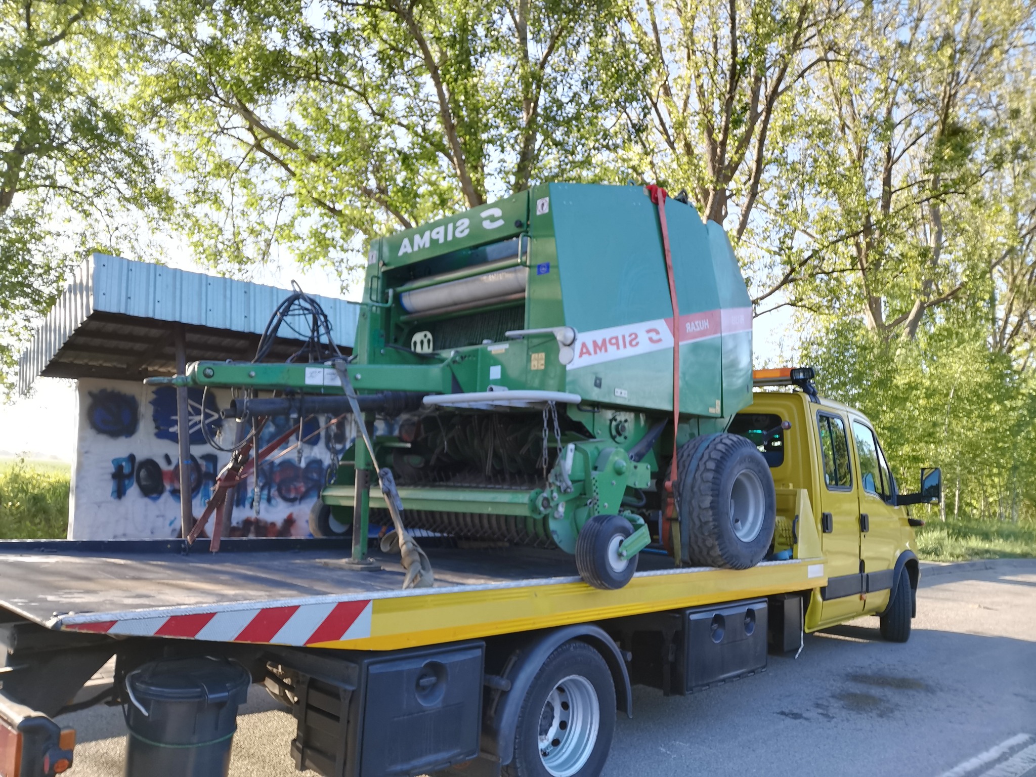 Żółta laweta przewozi zieloną prasę zwijającą SIPMA na tle drzew. Widoczny fragment budynku z graffiti. Ujęcie z dołu, podkreślające gabaryty maszyny rolniczej.