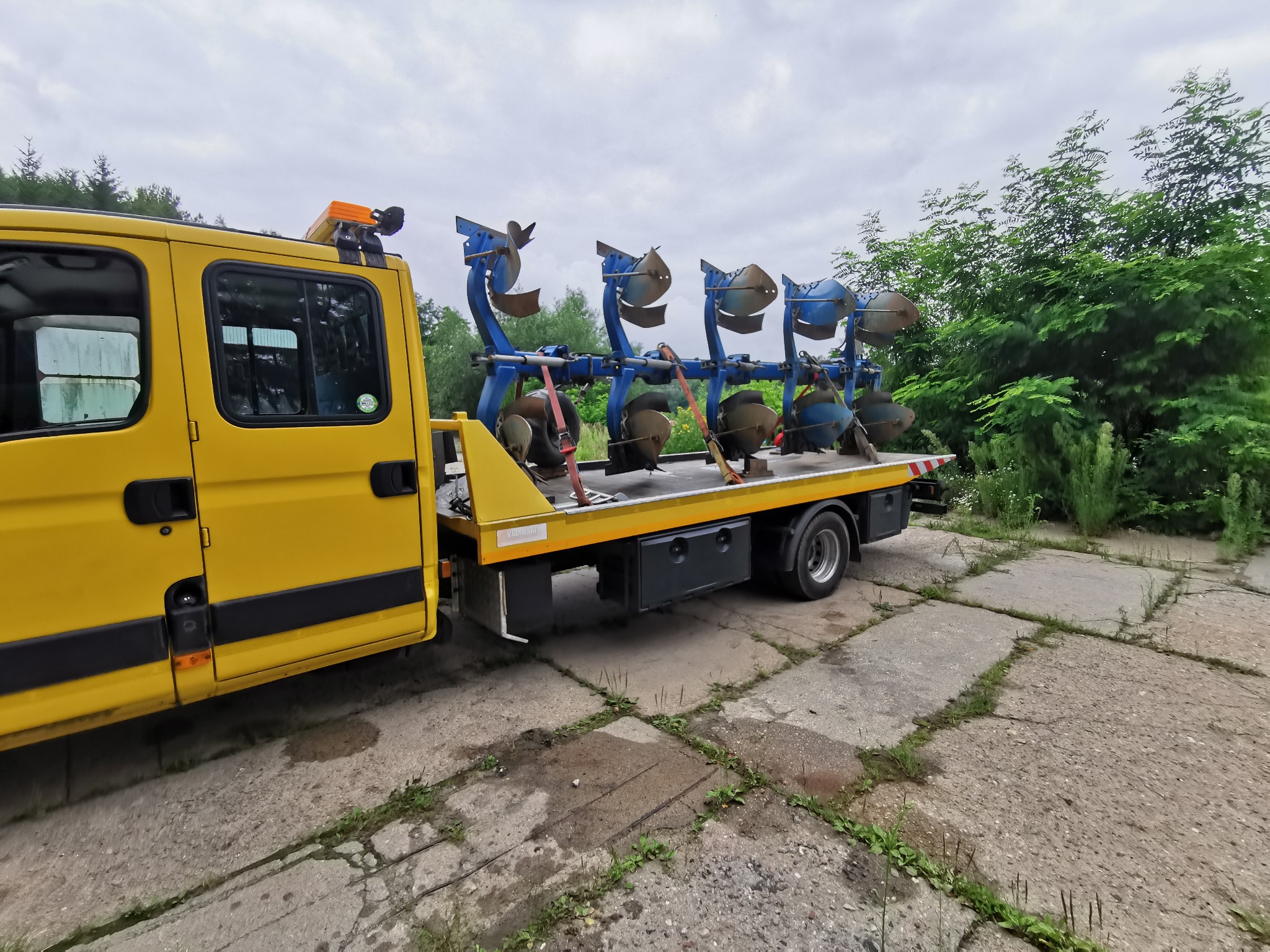Żółta laweta z niebieskim pługiem rolniczym na platformie, zabezpieczonym pasami transportowymi. Widok z boku na betonowym podłożu i zieleń w tle.