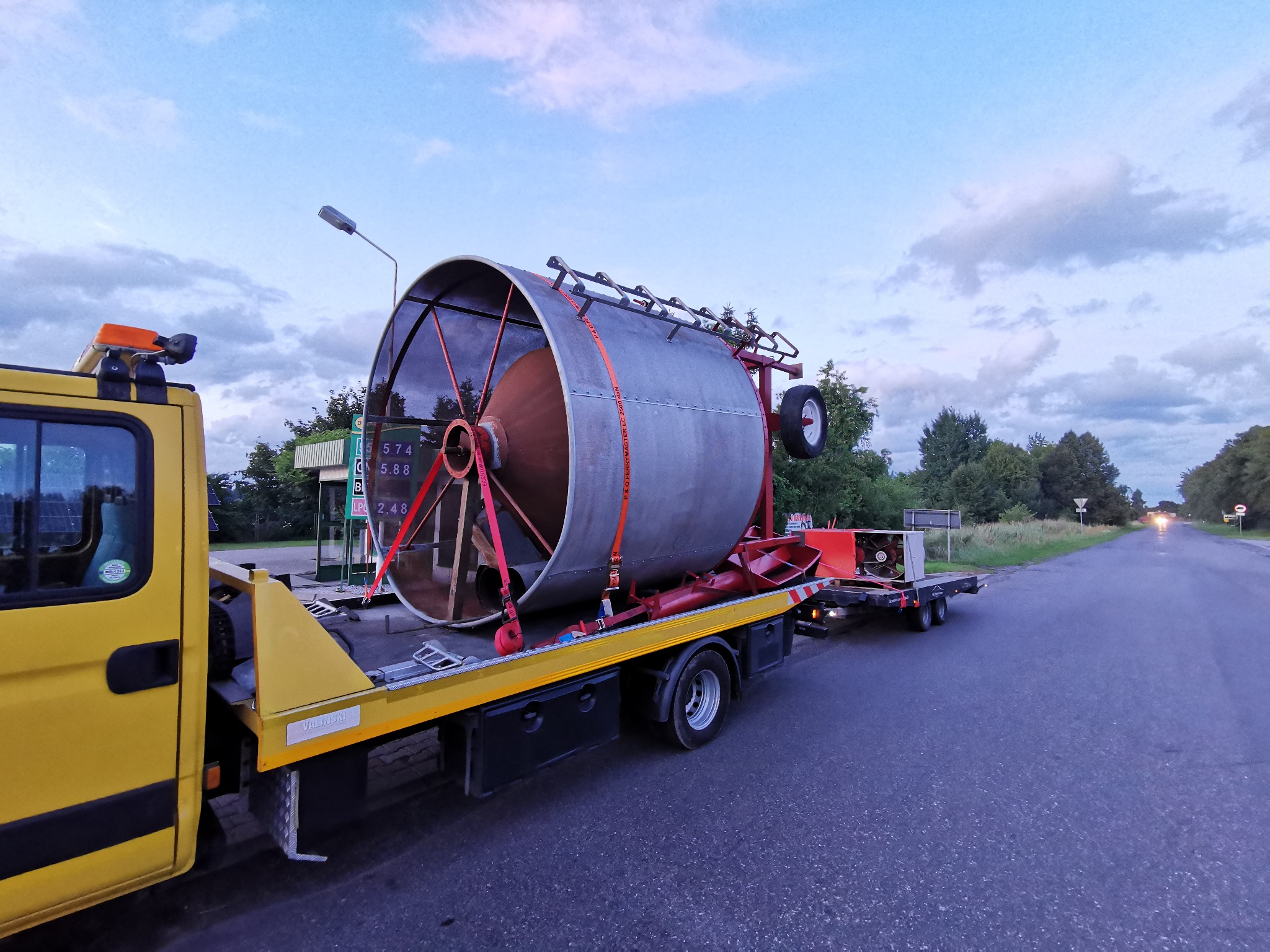 Żółta laweta z ładunkiem cylindrycznym zabezpieczonym pasami transportowymi na drodze w Złotowie. Widok z boku, w tle stacja paliw i zieleń.