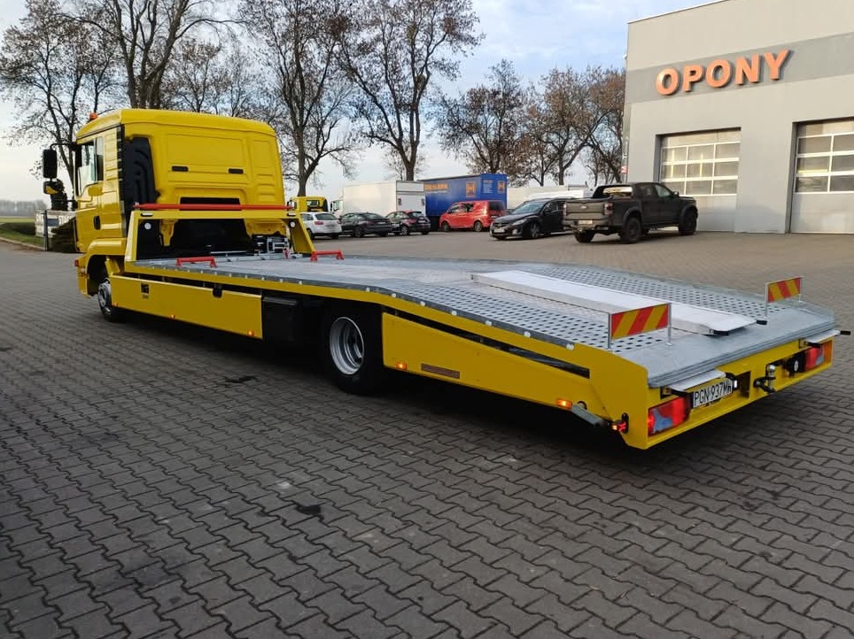 Żółta laweta z platformą załadunkową, zaparkowana na kostce brukowej. W tle budynek z napisem 'OPONY' i zaparkowane samochody. Widok z tyłu, lekko z boku.