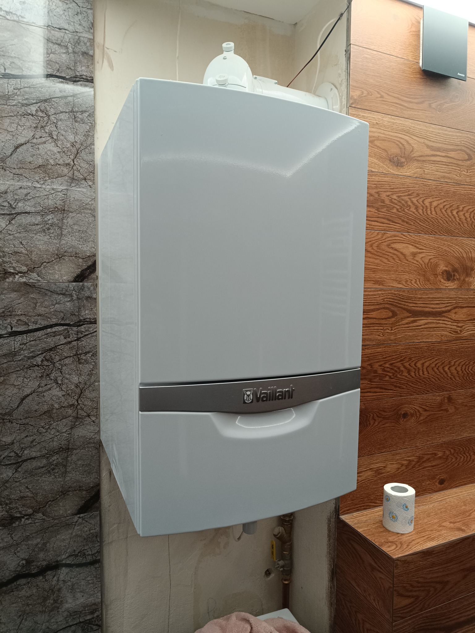 Biały piec gazowy Vaillant zamontowany na ścianie z marmurowymi i drewnianymi panelami. Widoczne rury instalacyjne i rolka papieru toaletowego.