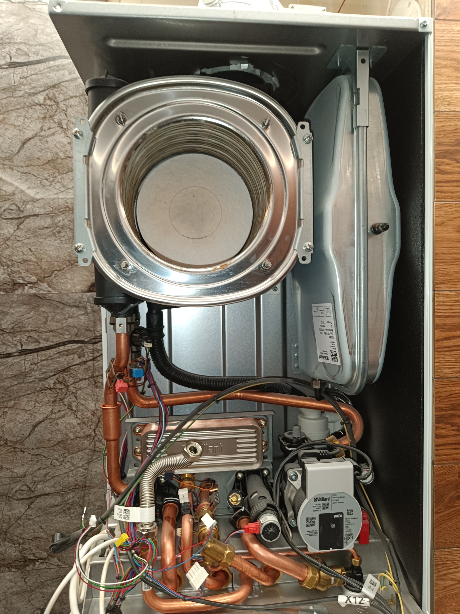 Otwarty piec gazowy Vaillant z widocznymi elementami wewnętrznymi, miedzianymi rurami i przewodami, na tle marmurowej i drewnianej ściany.