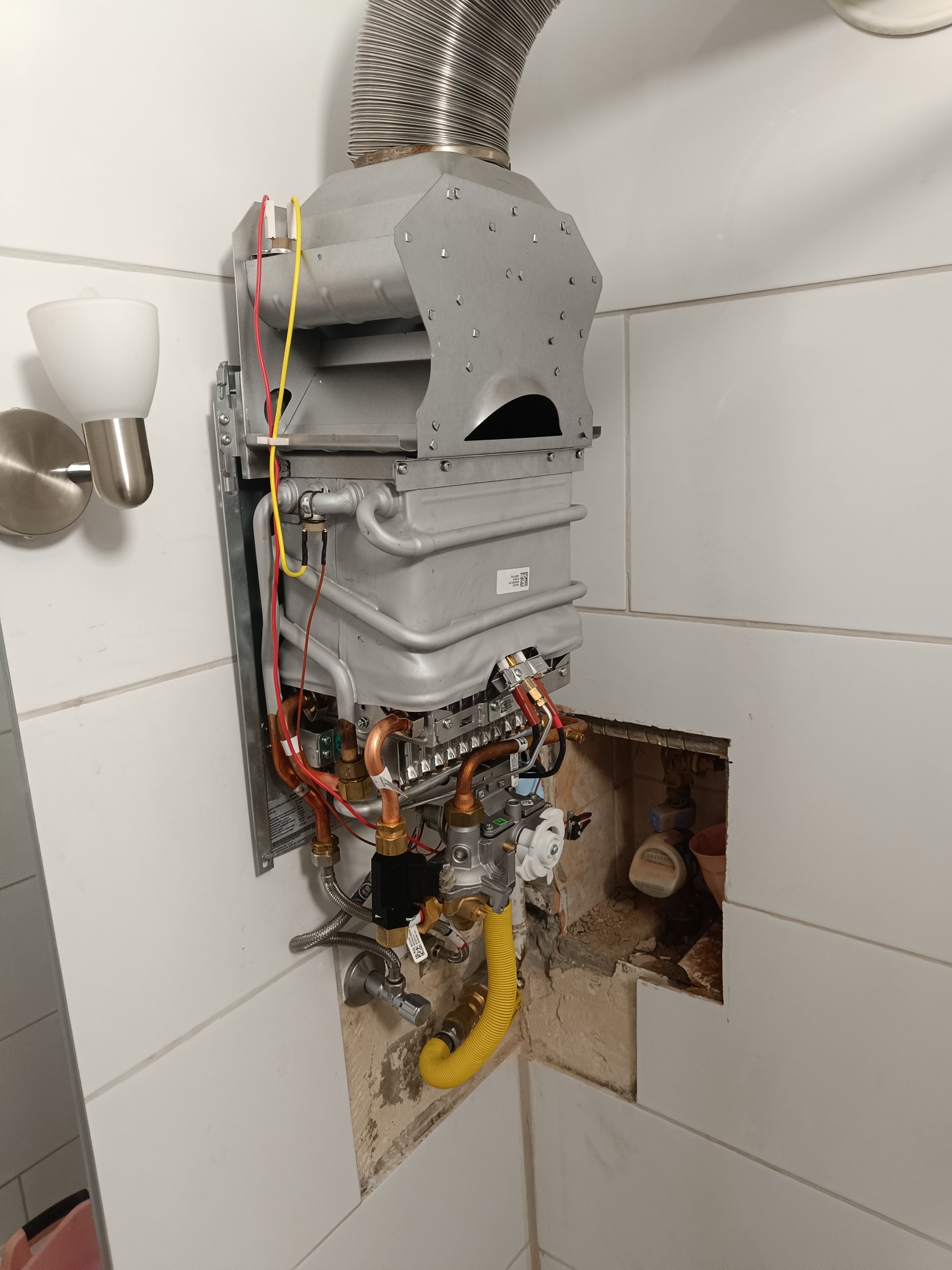 Szczegółowe ujęcie wiszącego pieca gazowego z podłączonymi przewodami, widoczne elementy instalacji w łazience z białymi płytkami i lampą ścienną.