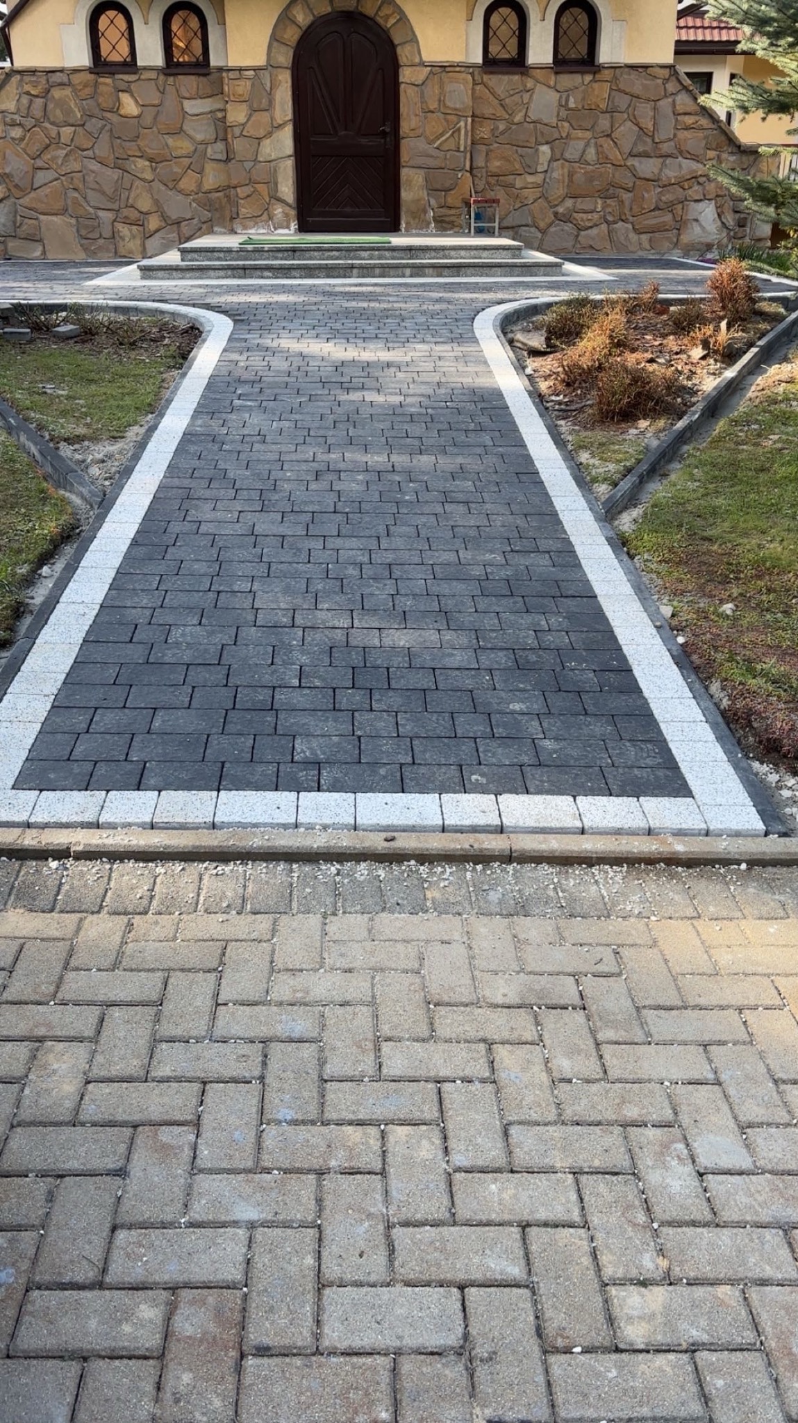 Ułożona ścieżka z ciemnej kostki brukowej obramowana jasną kostką, prowadząca do budynku z kamienną fasadą i brązowymi drzwiami. Widok z perspektywy.