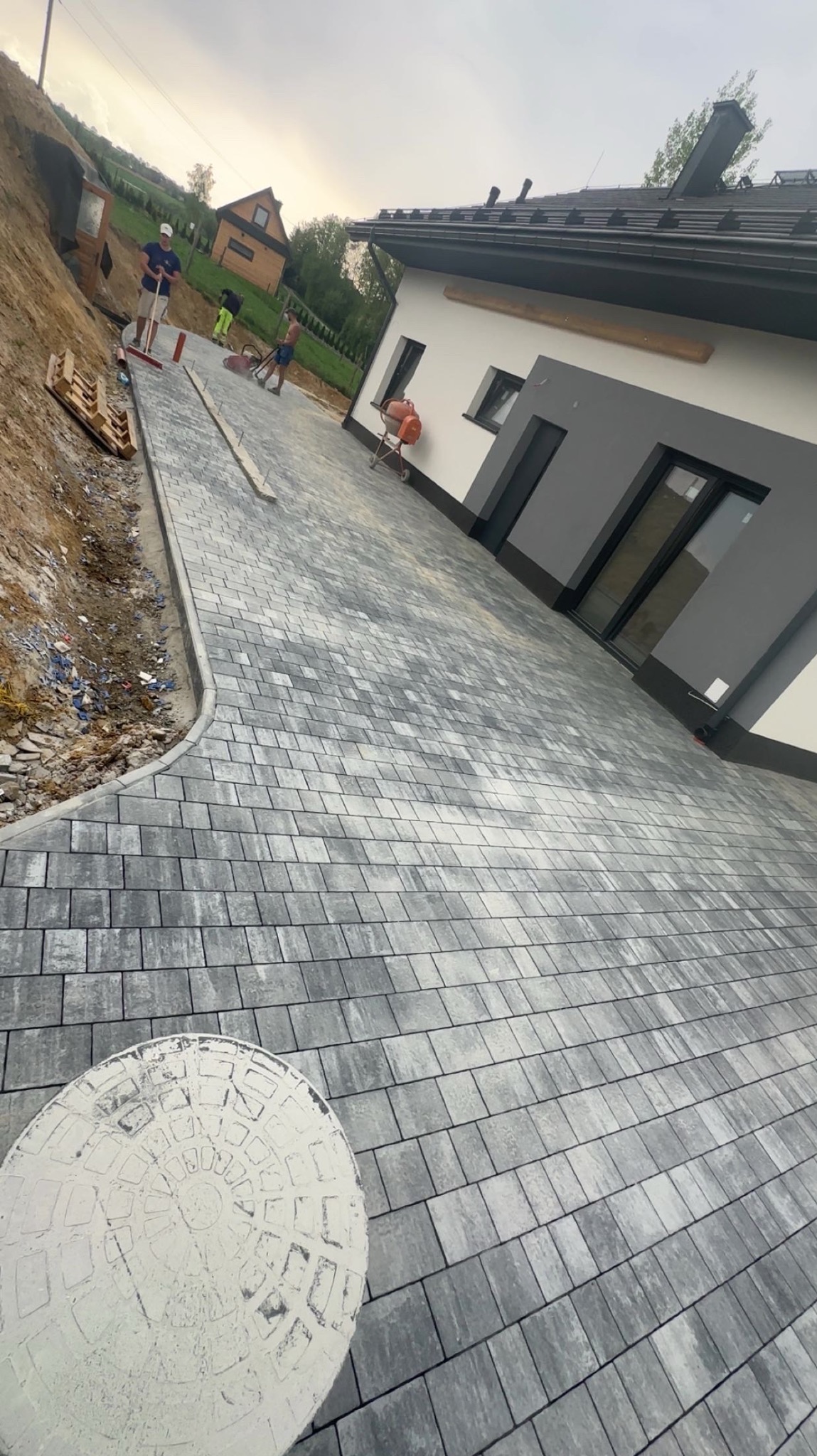 Ułożona szara kostka brukowa na podjeździe domu. W tle pracownicy, betoniarka i paleta. Widoczny właz kanalizacyjny.