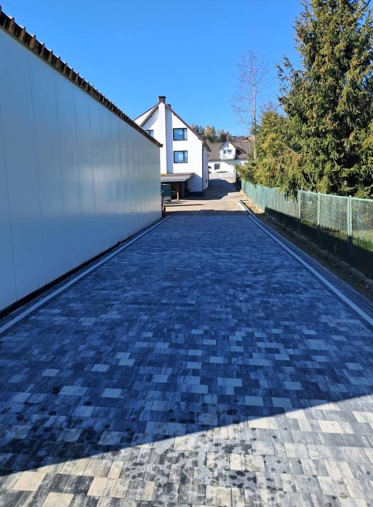 Nawierzchnia z szarej kostki brukowej ułożona wzdłuż ścieżki między budynkami. Widoczne białe obrzeża i zielone ogrodzenie w tle.