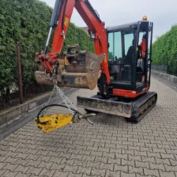Pomarańczowa mini koparka Kubota U27-4 z młotem hydraulicznym zawieszonym na łańcuchach, stojąca na kostce brukowej, na tle zielonego żywopłotu.