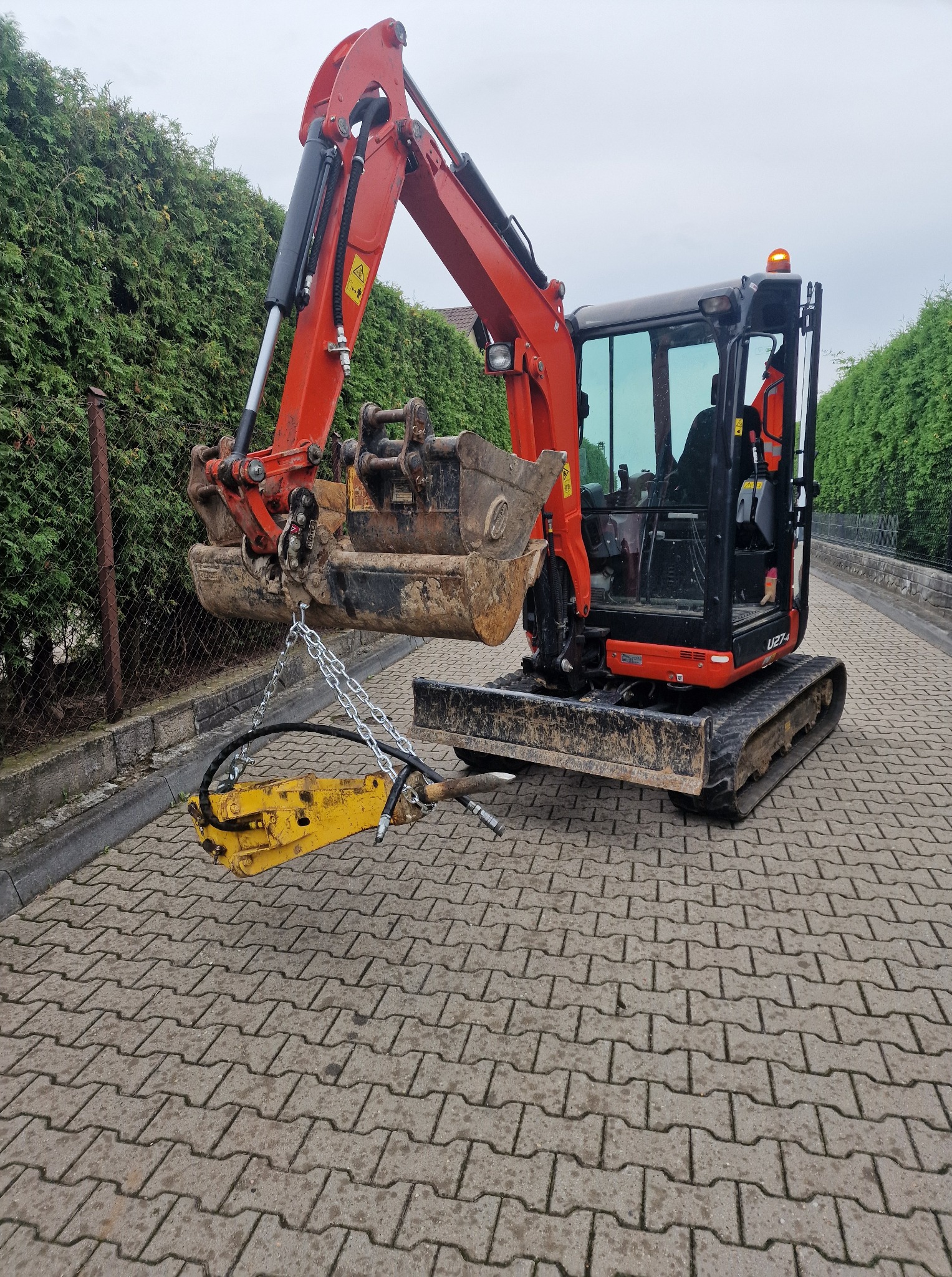 Pomarańczowa mini koparka Kubota U27-4 z młotem hydraulicznym zawieszonym na łańcuchach, stojąca na kostce brukowej, na tle zielonego żywopłotu.