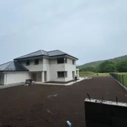 Nowoczesny dom z wyrównanym terenem pod trawnik. Osoba z linką wyznacza granice. W tle zalesione wzgórze i pochmurne niebo.