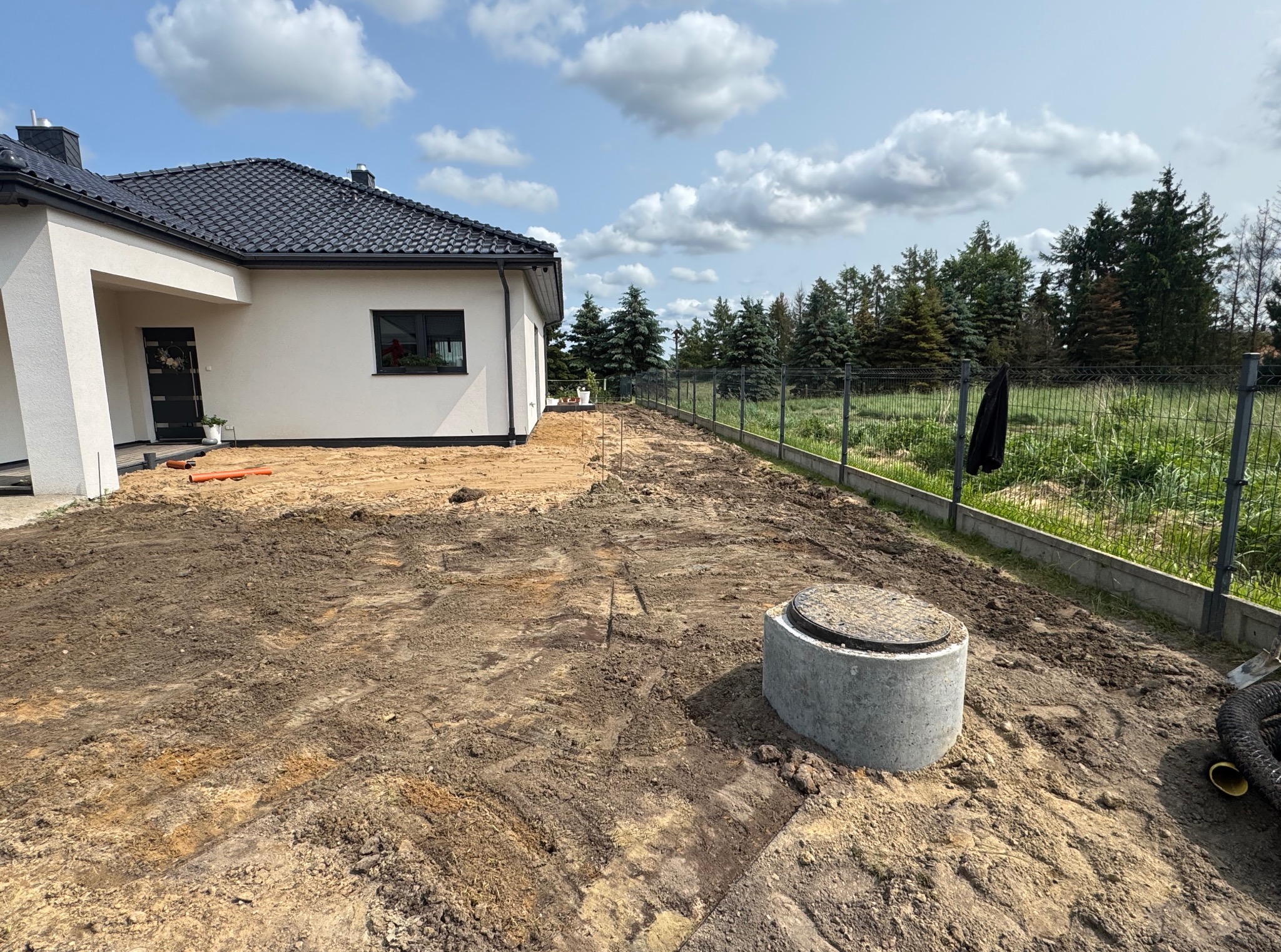 Przygotowany teren pod budowę domu jednorodzinnego z widocznym betonowym kręgiem szamba i wykopanymi rowami pod instalacje. Słoneczny dzień, błękitne niebo z chmurami.