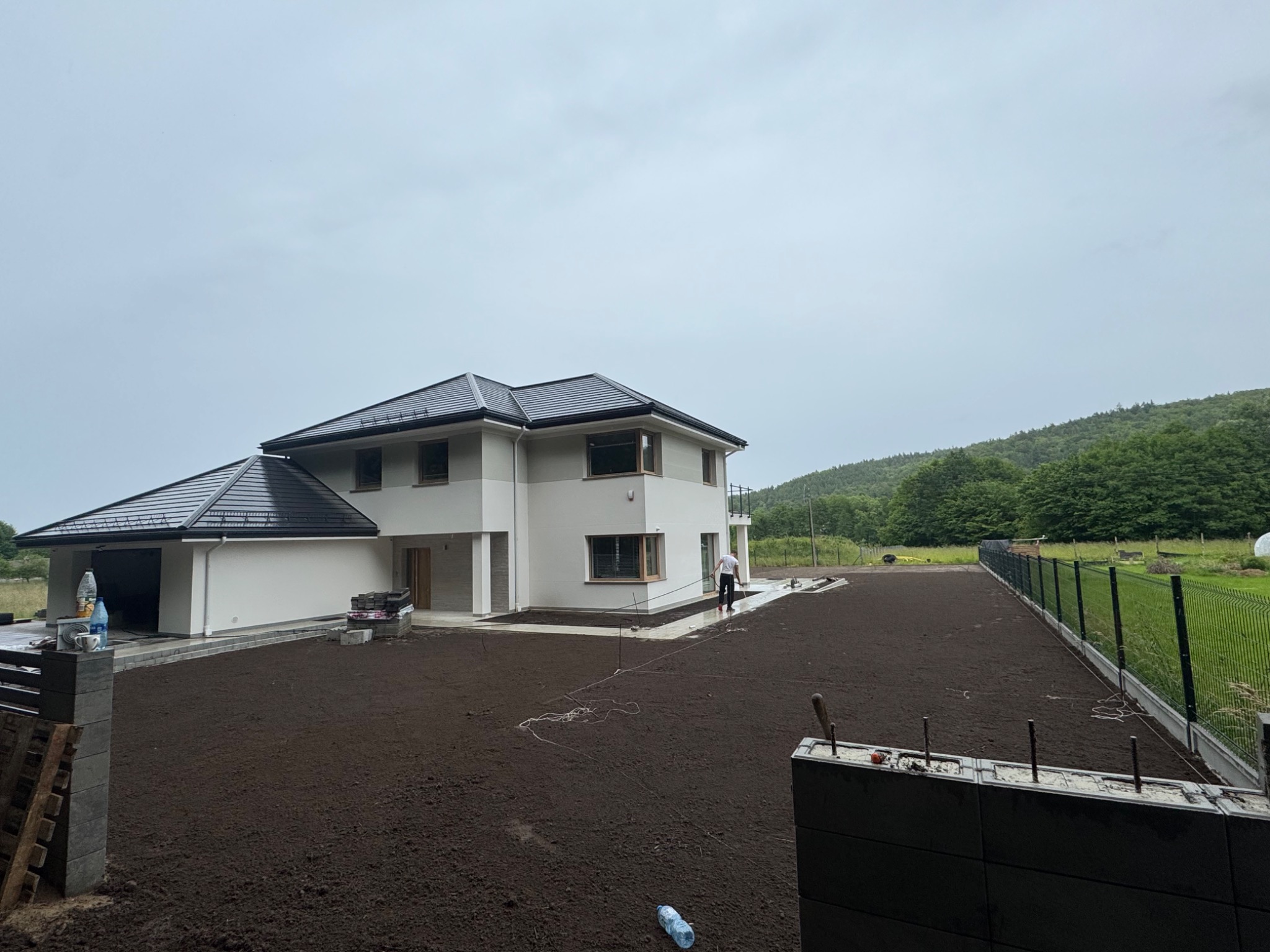 Nowoczesny dom z wyrównanym terenem pod trawnik. Osoba z linką wyznacza granice. W tle zalesione wzgórze i pochmurne niebo.