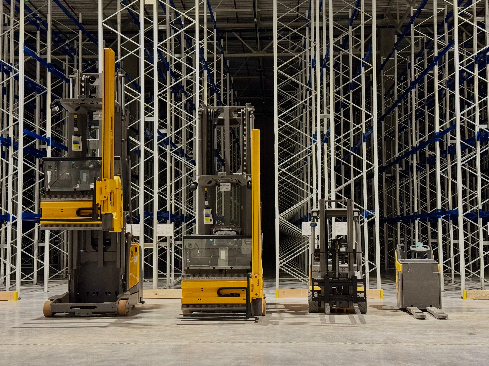 Cztery wózki widłowe Jungheinrich w magazynie wysokiego składowania z regałami paletowymi. Wózki różnią się wielkością i typem, od wózka wysokiego składowania do wózka paletowego.