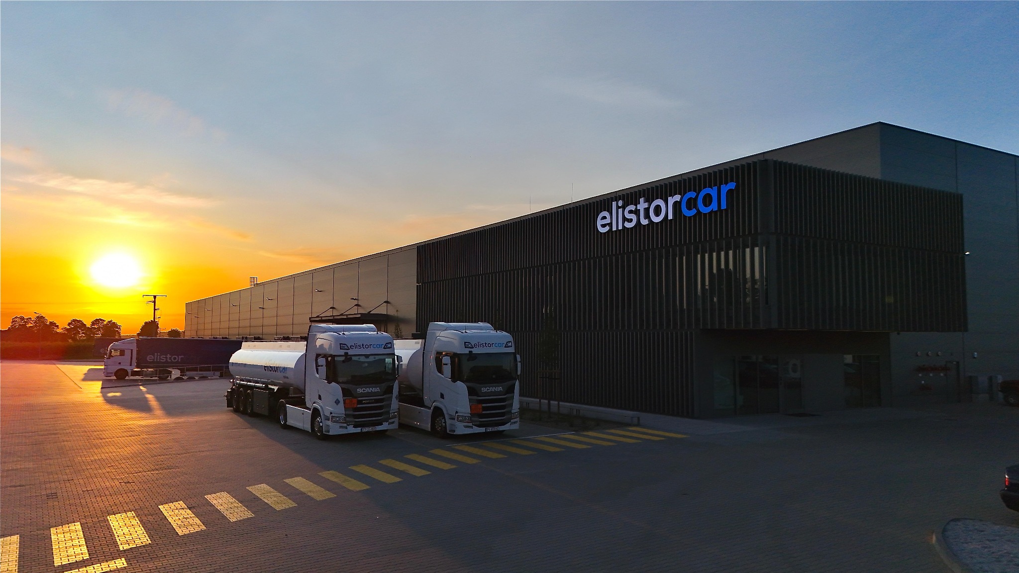 Dwa białe ciągniki siodłowe z cysternami marki Scania na tle nowoczesnego magazynu z logo 'elistorcar' i zachodzącego słońca.