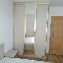 WEST ART Pracownia Meblarska - Jasna szafa przesuwna z lustrem w sypialni, widok na wnętrze pokoju odbijający się w lustrzanych drzwiach, obok drewniane drzwi.