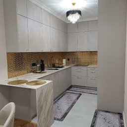 WEST ART Pracownia Meblarska - Jasna kuchnia z złotymi akcentami: mozaika nad blatem, złota armatura i dekoracje. Nowoczesne oświetlenie i dywaniki dopełniają elegancki wystrój.