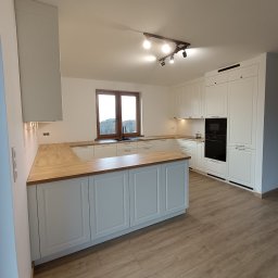 WEST ART Pracownia Meblarska - Jasna kuchnia z drewnianym blatem, wyspą i oknem. Uwagę przykuwają czarne, minimalistyczne uchwyty szafek i lampa sufitowa z reflektorami. Parkiet w odcieniach brązu.