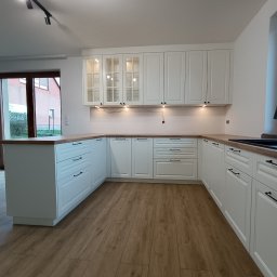 WEST ART Pracownia Meblarska - Jasna kuchnia w stylu klasycznym z drewnianym blatem i frezowanymi frontami. Górne szafki częściowo przeszklone, oświetlenie punktowe. Podłoga z paneli imitujących drewno.