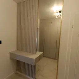 Nowoczesna zabudowa przedpokoju z lustrem w złotej ramie i konsolą z lamelami w odcieniach beżu. Widoczne szafy w tle i marmurowa posadzka.