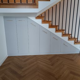 WEST ART Pracownia Meblarska - Szafa wnękowa pod schodami z szarymi frontami i złotymi uchwytami, parkiet w jodełkę. Zabudowa wnęki pod schodami.