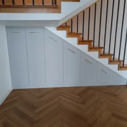 Szafa wnękowa pod schodami z szarymi frontami i złotymi uchwytami, parkiet w jodełkę. Zabudowa wnęki pod schodami.