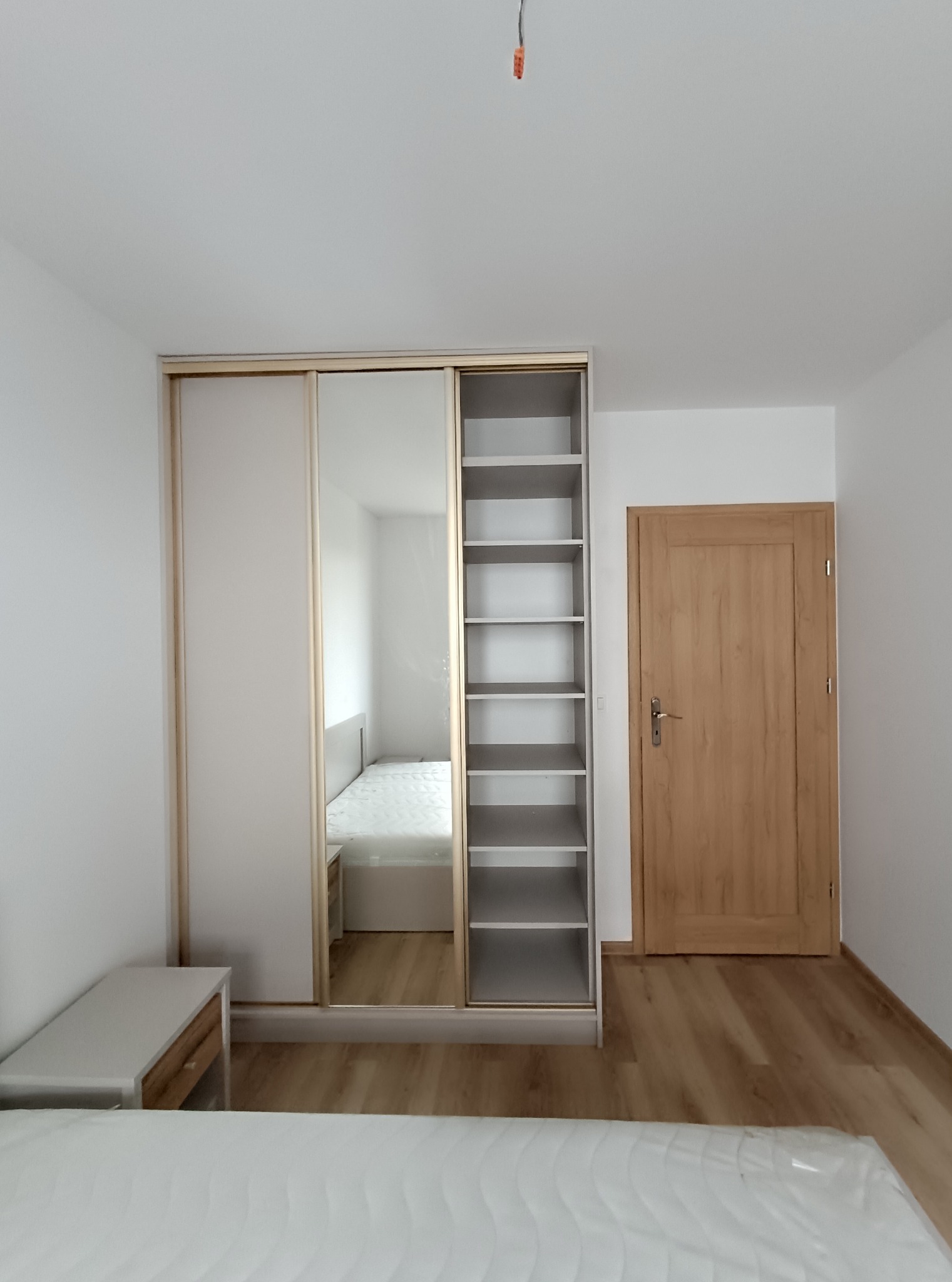 Jasna szafa wnękowa z przesuwnymi drzwiami i lustrem w pokoju. Widoczny fragment łóżka i szafka nocna. Minimalistyczny design, drewniana podłoga.