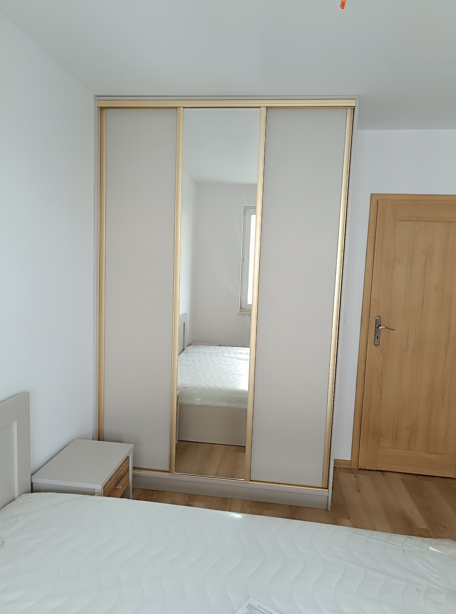 Jasna szafa przesuwna z lustrem w sypialni, widok na wnętrze pokoju odbijający się w lustrzanych drzwiach, obok drewniane drzwi.