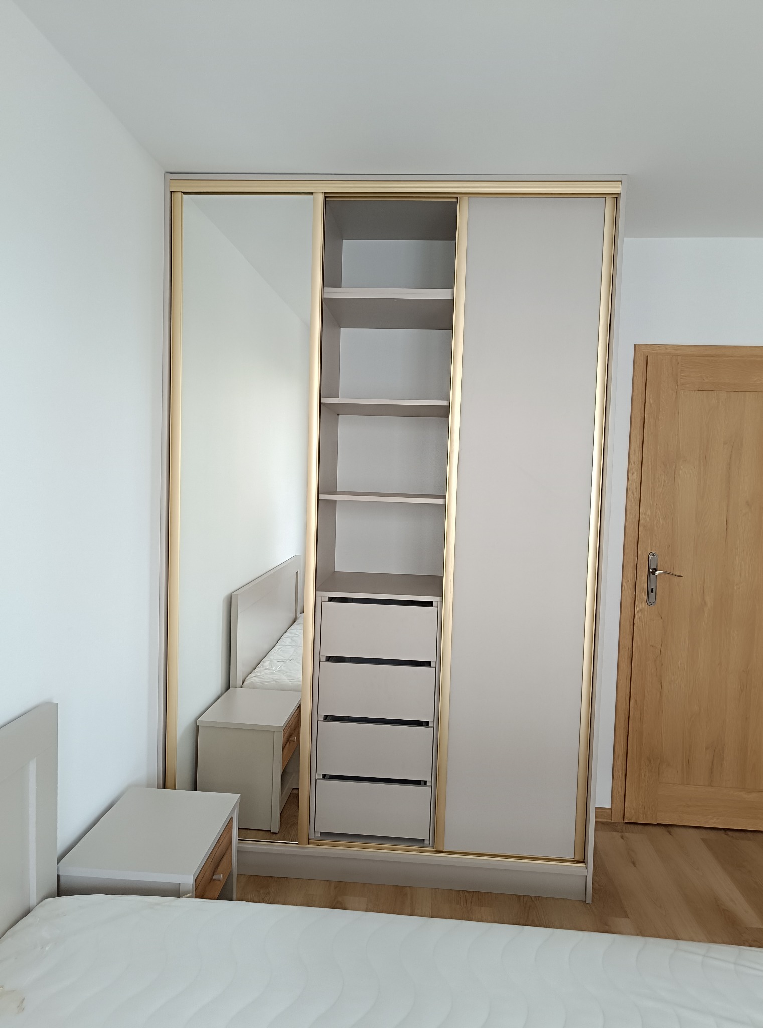 Jasna szafa przesuwna z lustrem i złotymi obramowaniami, wkomponowana w aranżację sypialni z widocznym łóżkiem i szafką nocną.