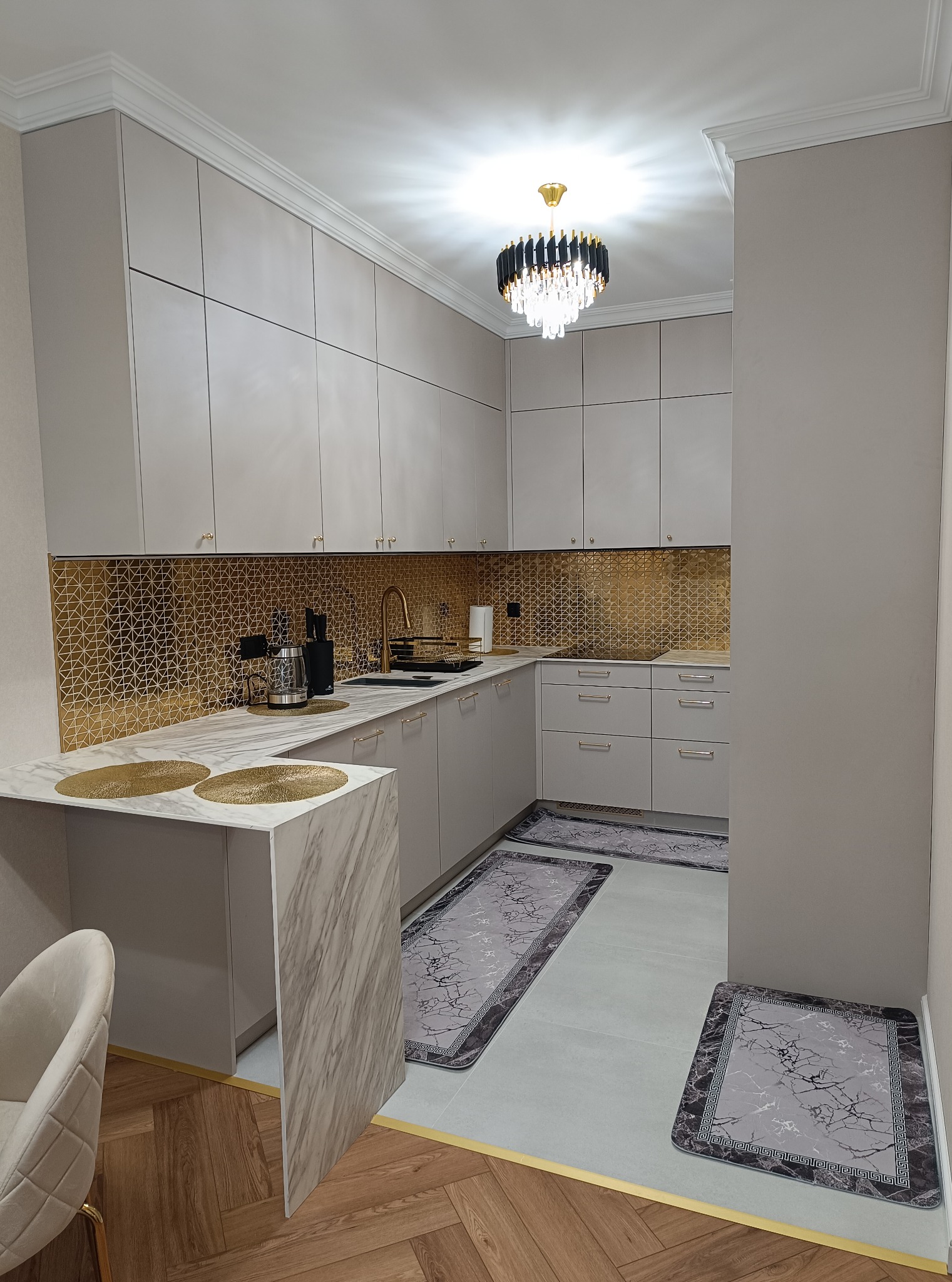 Jasna kuchnia z złotymi akcentami: mozaika nad blatem, złota armatura i dekoracje. Nowoczesne oświetlenie i dywaniki dopełniają elegancki wystrój.