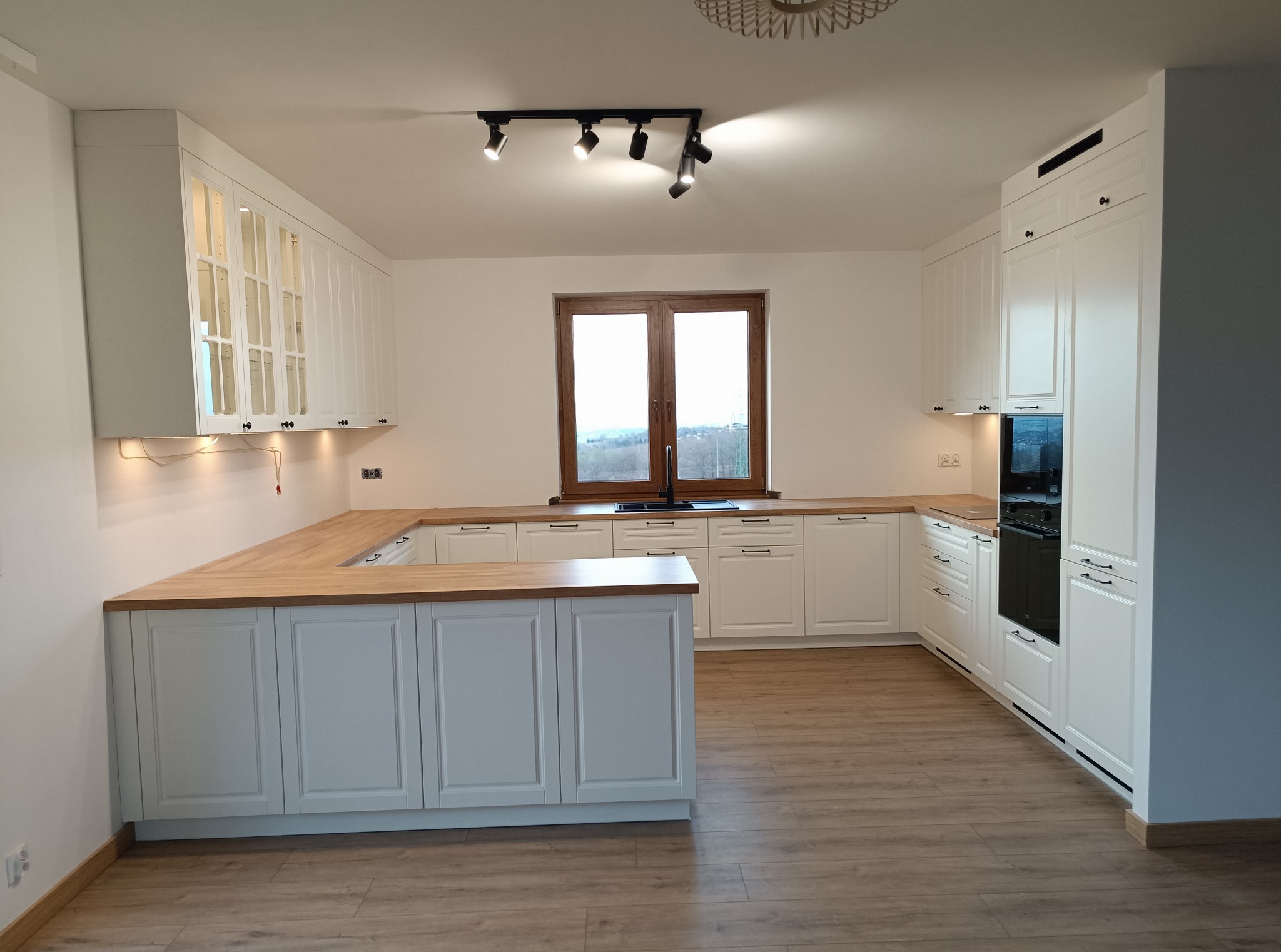 Jasna kuchnia w stylu klasycznym z drewnianym blatem. Białe szafki z ciemnymi uchwytami. Na podłodze panele imitujące drewno. Okno z widokiem na zewnątrz.