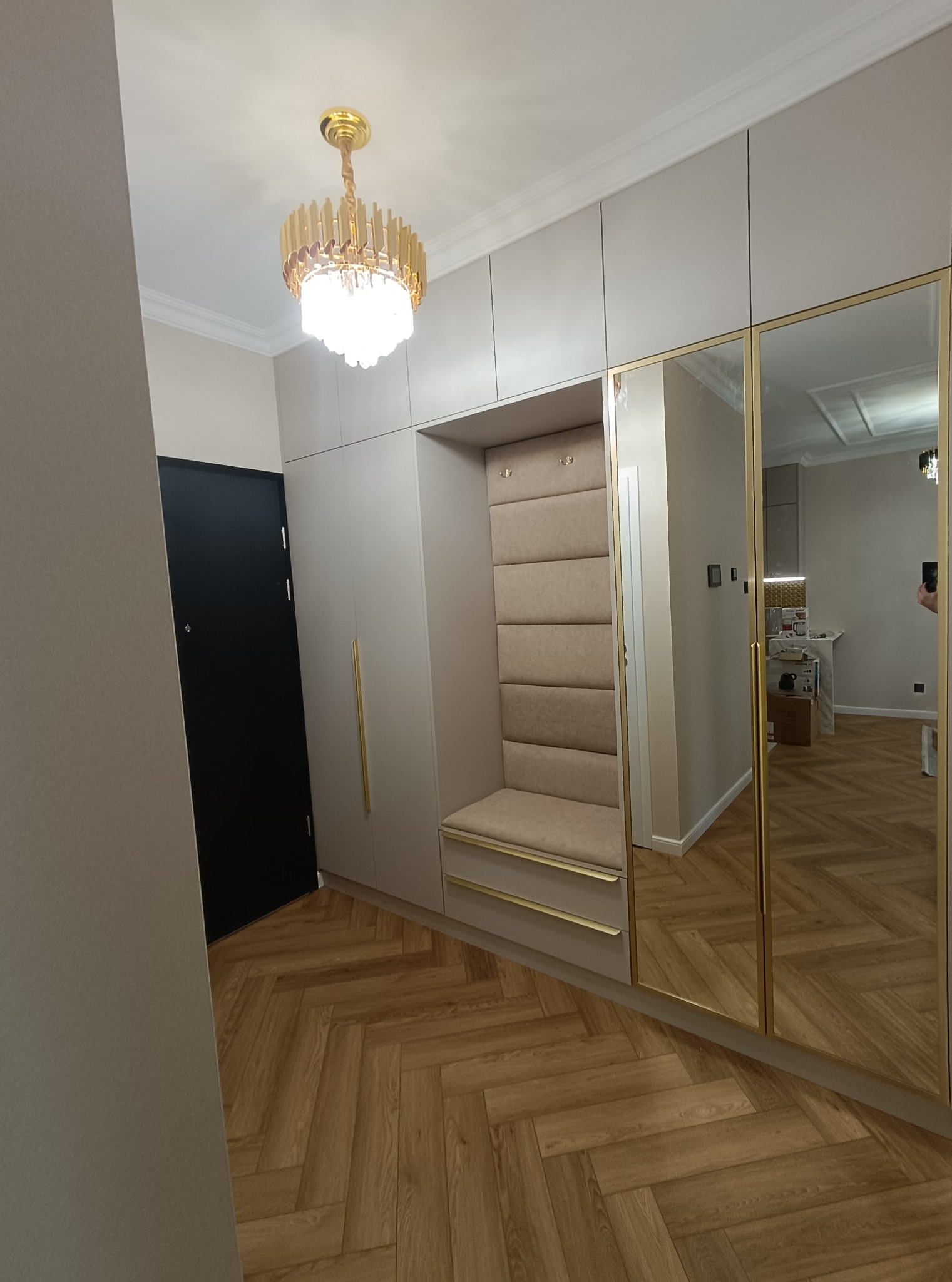 Beżowa szafa wnękowa z siedziskiem i lustrzanymi drzwiami, złote akcenty, parkiet w jodełkę. Nowoczesne oświetlenie sufitowe. Widok na przedpokój i fragment kuchni w odbiciu.